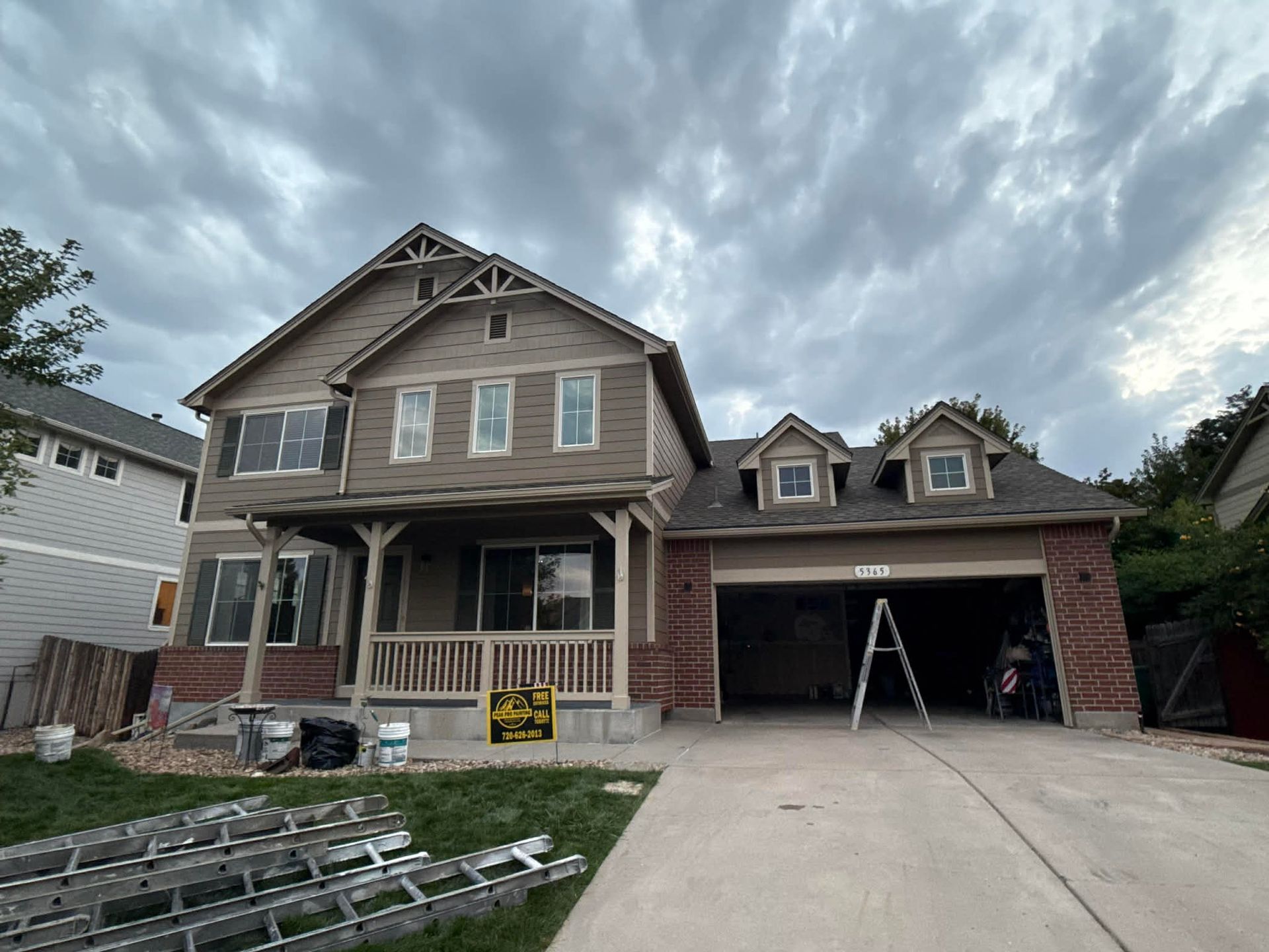A two-story tan house with a brick base and a front porch, featuring a wide garage and ladders resting on the lawn.