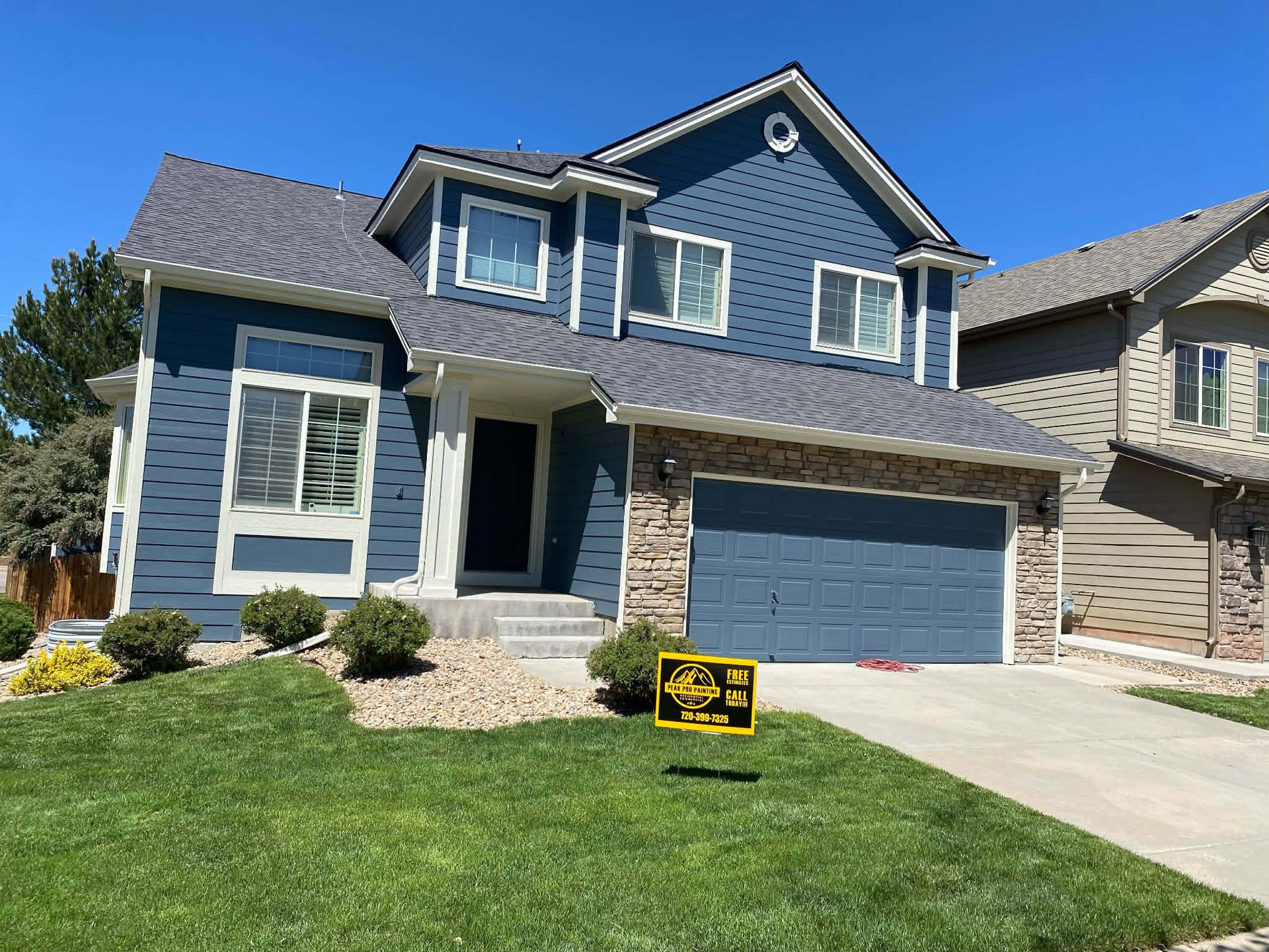 A two-story blue house with stone accents, a garage, and a yellow sign on the lawn under a clear blue sky.