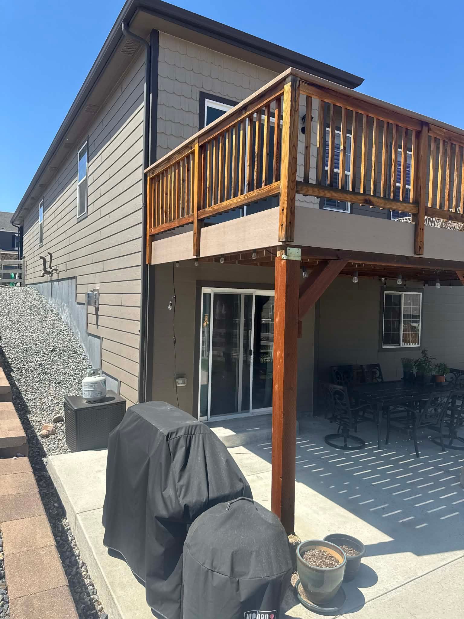 A light brown house with a wooden deck, a concrete patio below featuring a grill, and a gravel slope along the side.