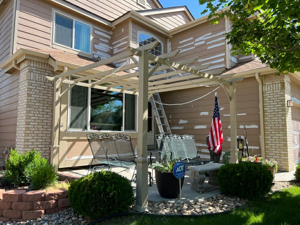 A tan house with a beige pergola on a patio, featuring a small American flag and a ladder leaning against the siding.