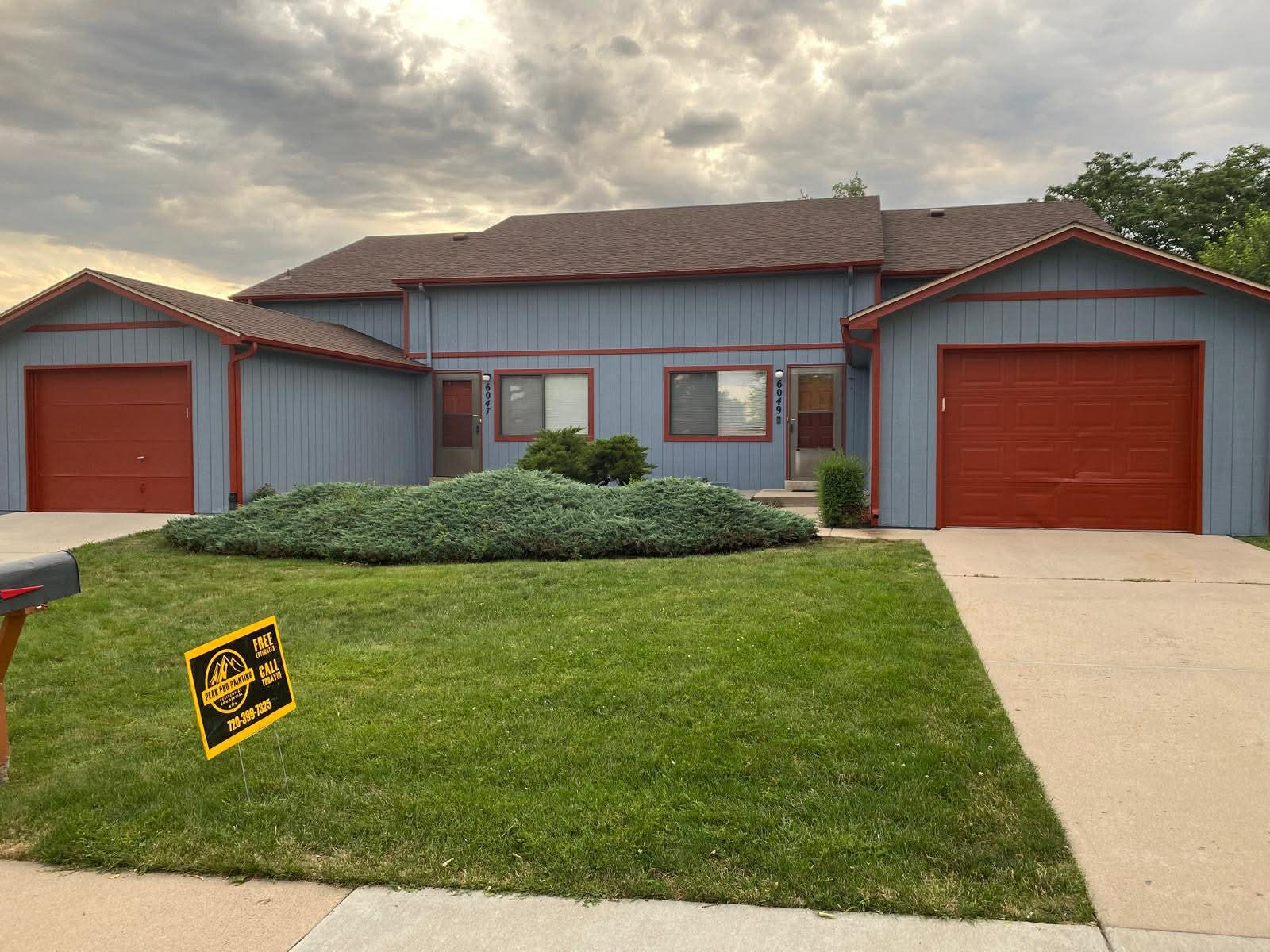 A blue two-story house with two orange garage doors, a brown roof, and a green lawn under a cloudy sky.