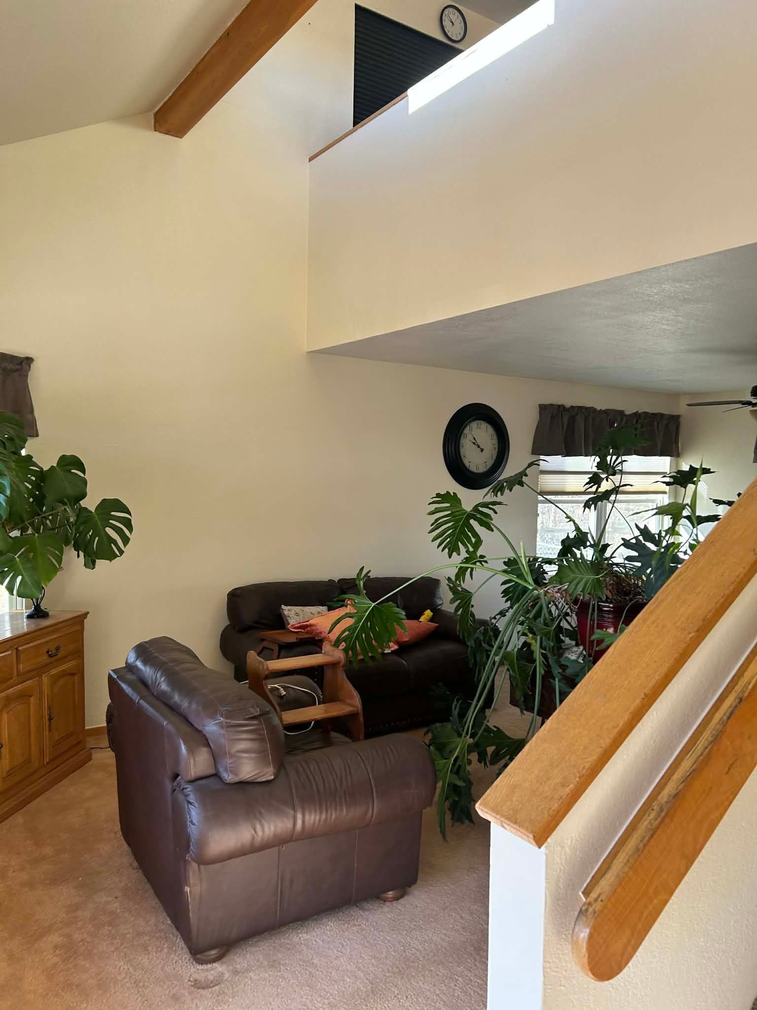 A living room with brown leather furniture, a wooden side table, large leafy plants, and a staircase with a wooden rail.