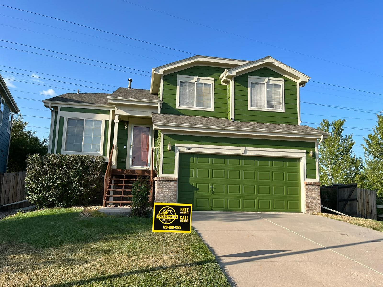 A two-story, olive-green house with a front-facing garage, tan trim, and a small front yard with a real estate sign.