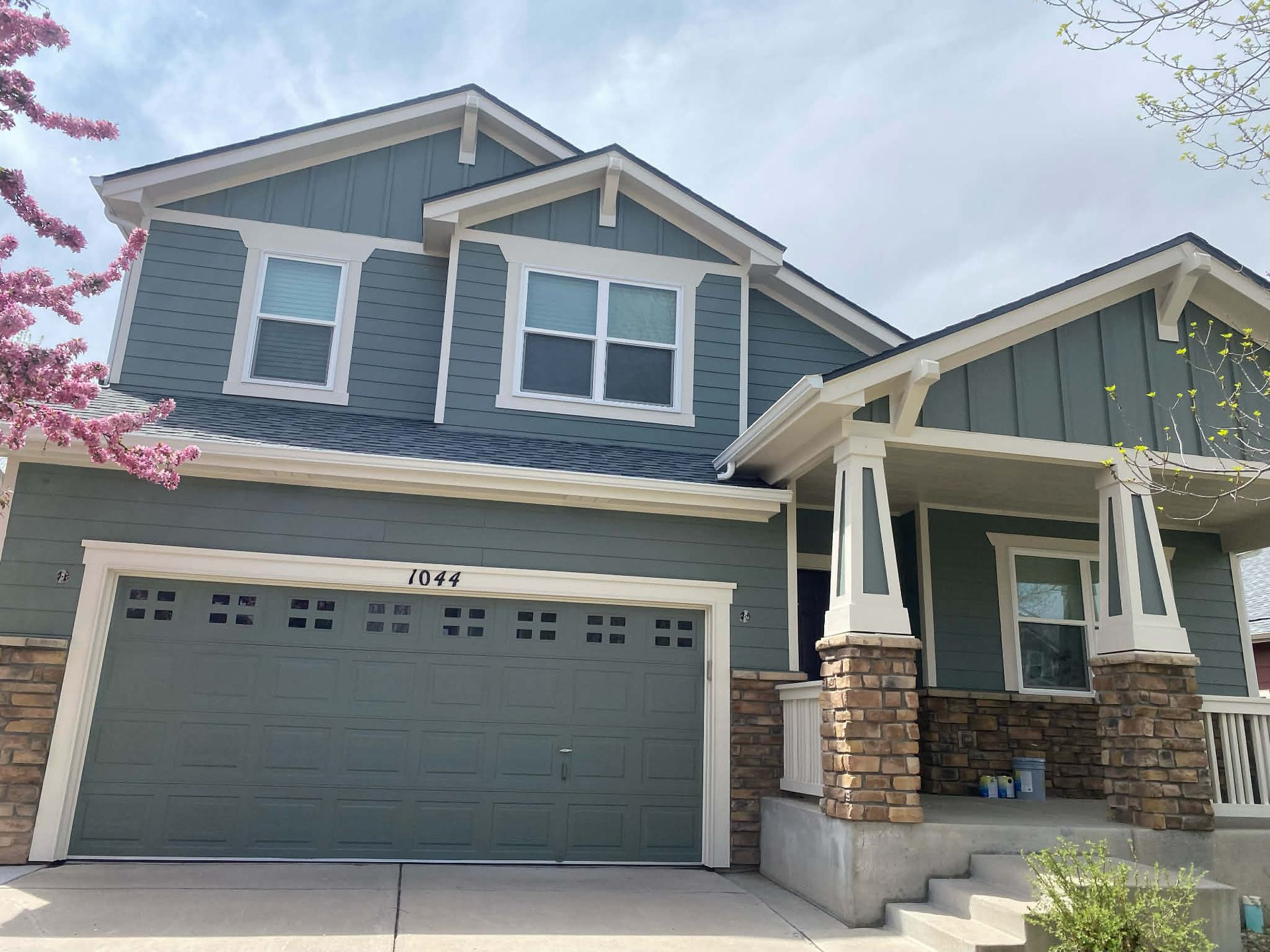 Two-story, blue-gray home with a two-car garage, stone masonry accents, a front porch, and a flowering tree on the left.