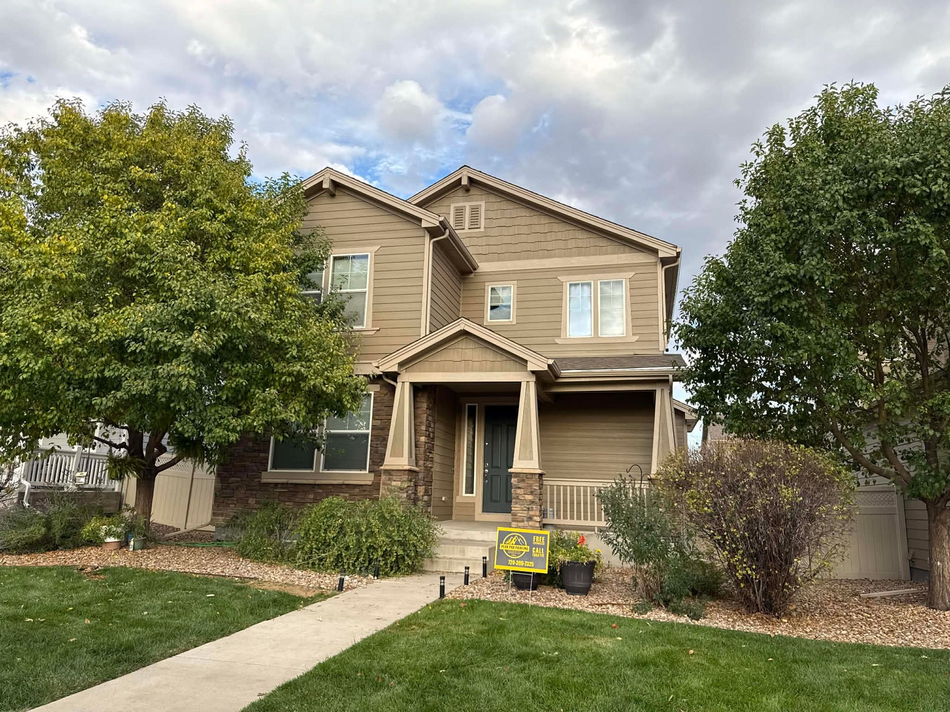 A two-story, tan suburban home with a stone-accented entryway, front yard, and trees under a cloudy sky.