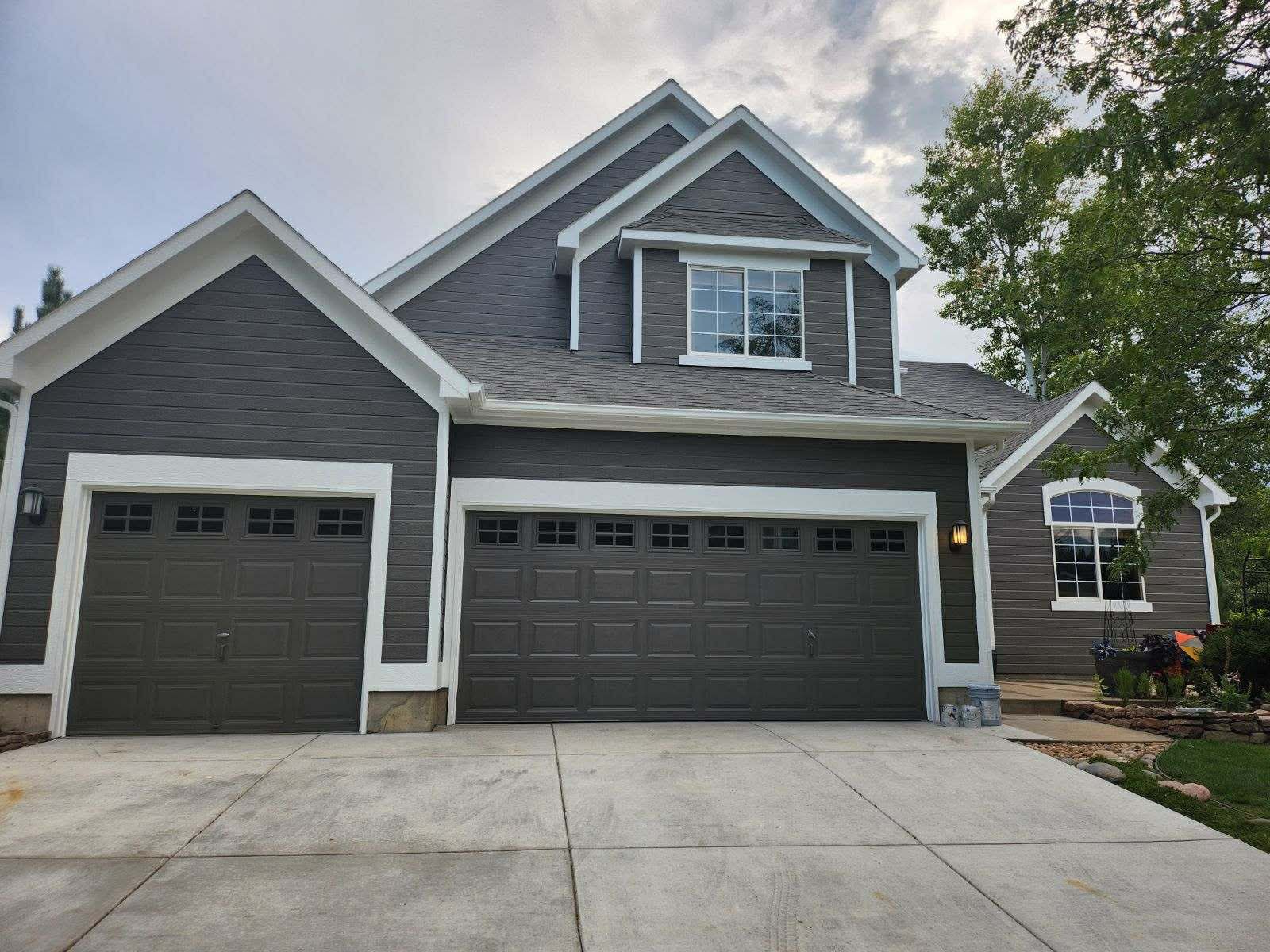 A two-story house with dark gray horizontal siding, a white trim, and two garage doors, viewed from the driveway.