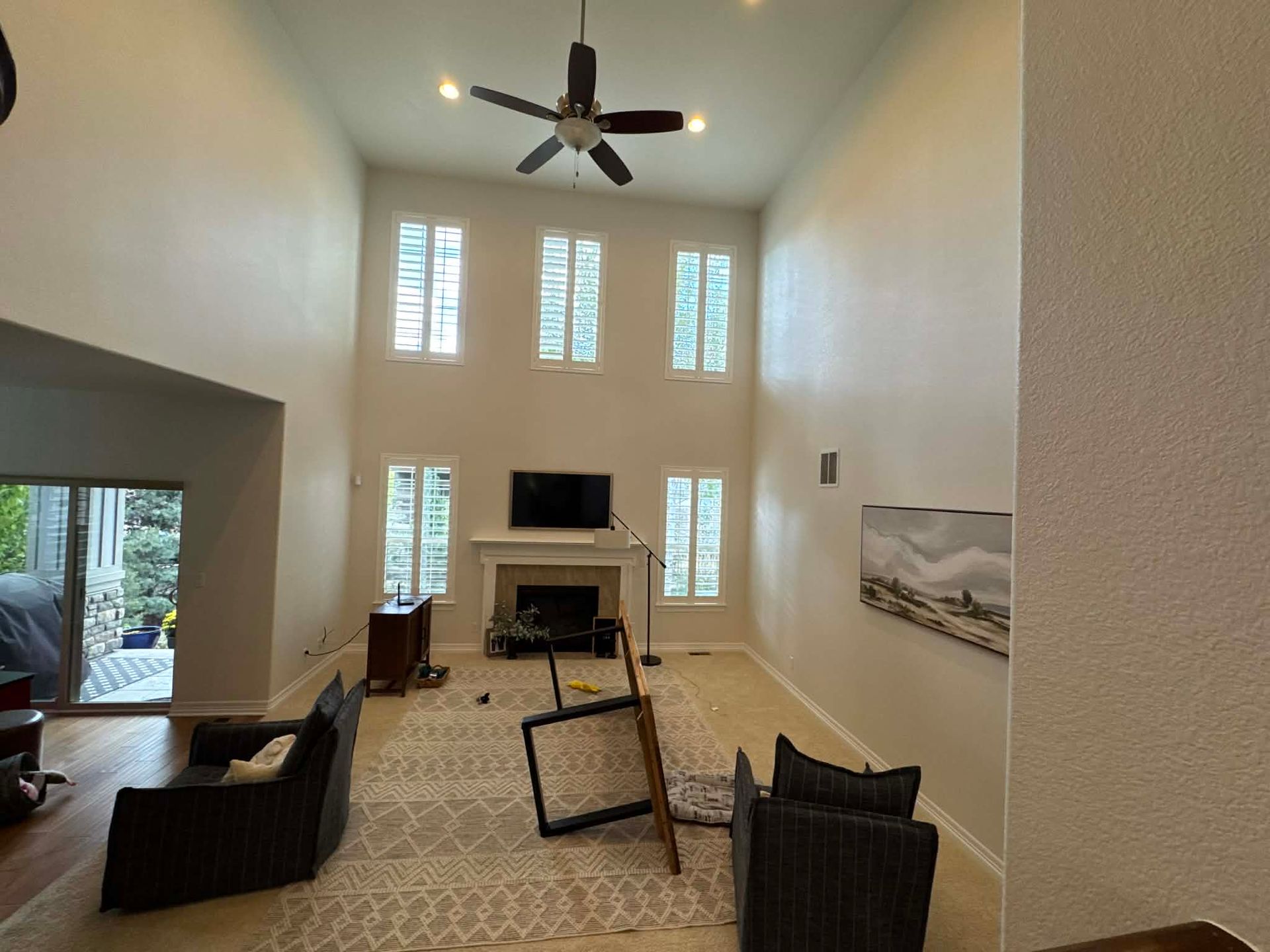 A spacious living room with a fireplace, tall windows, ceiling fan, carpet, and two armchairs facing a central table.