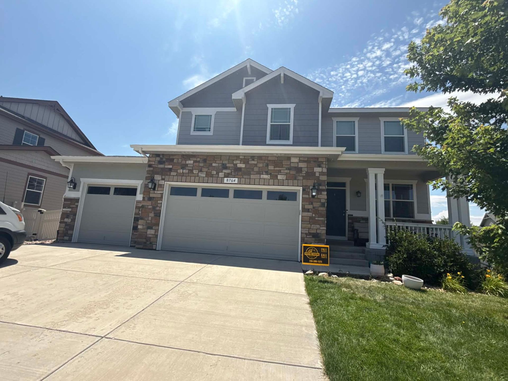 A two-story grey suburban house with stone accents, a three-car garage, and a front lawn under a blue sky.