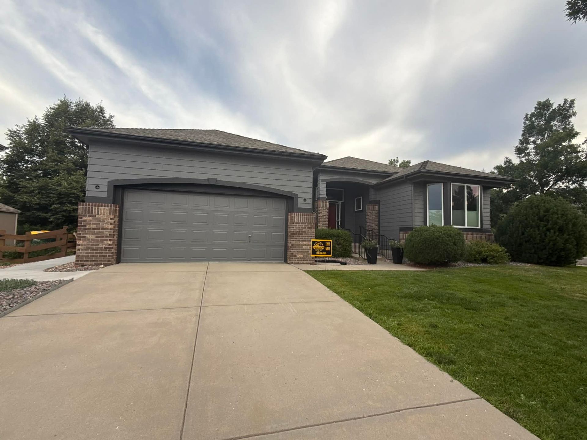 A single-story grey house with a two-car garage, stone trim, and a manicured lawn under a cloudy sky.