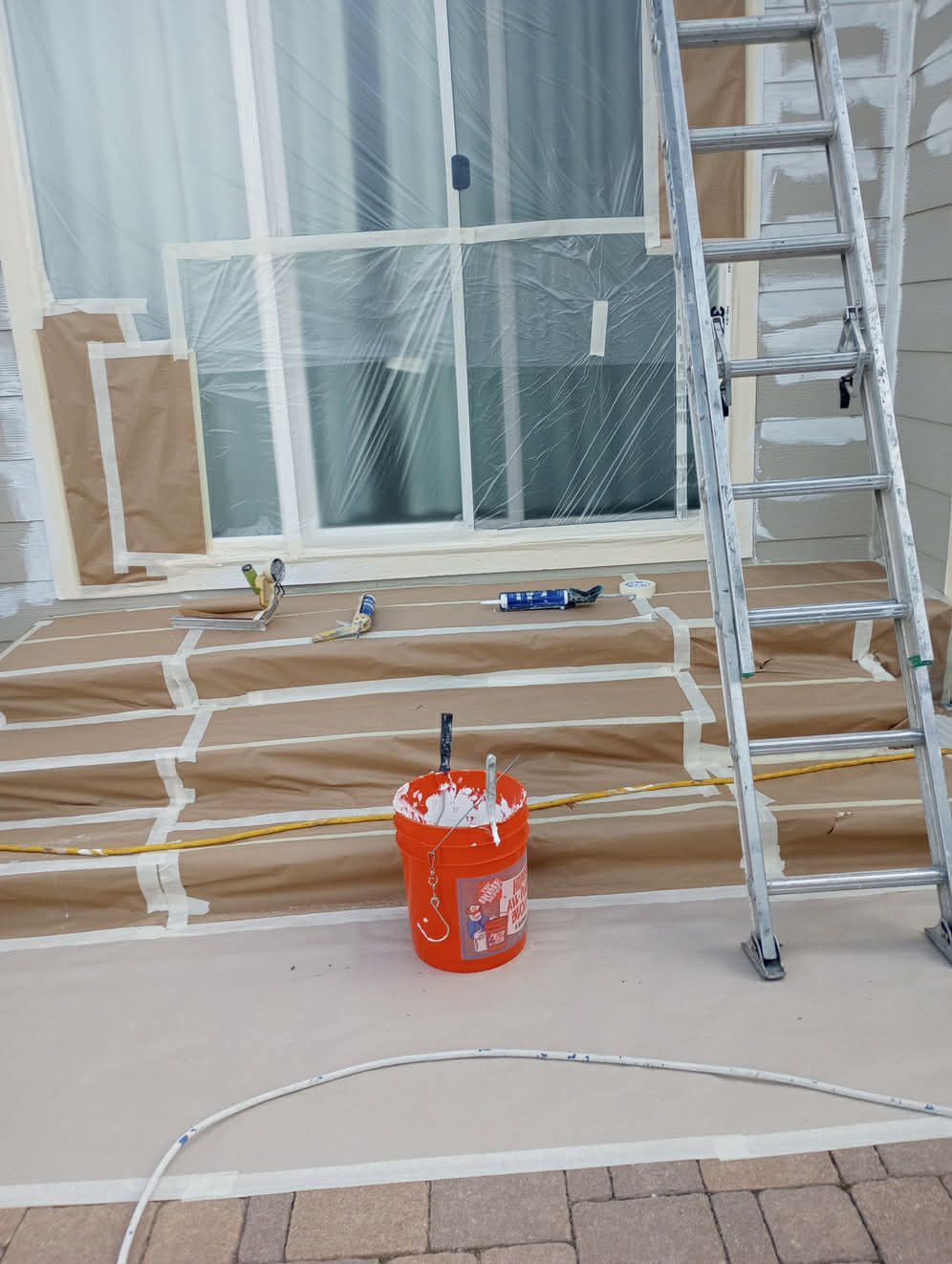 A patio under construction, with taped-off glass doors, paper-covered steps, a paint bucket, and a metal ladder.