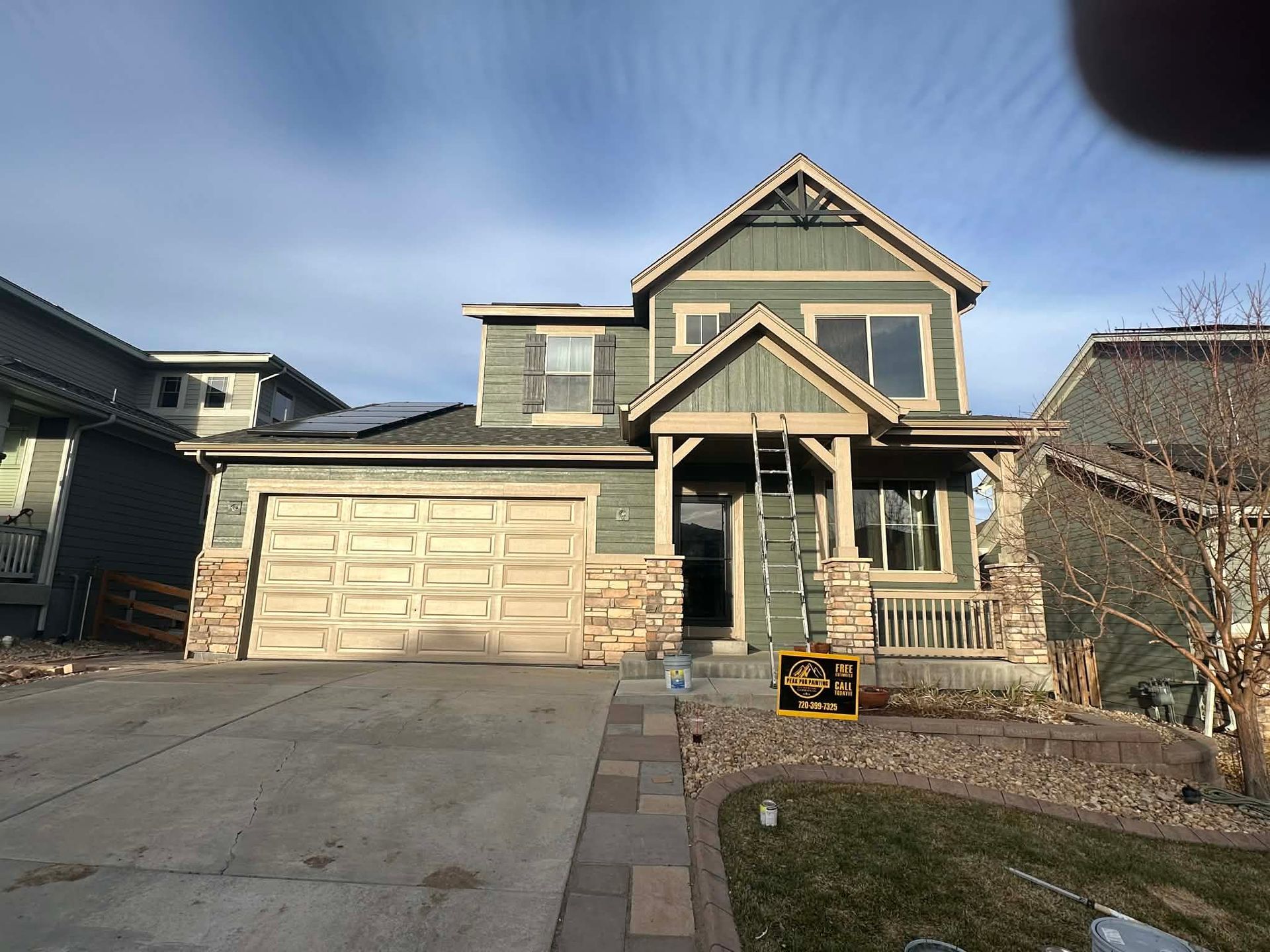 A two-story green suburban house with a tan garage, stone accents, and a front porch under a clear, bright blue sky.
