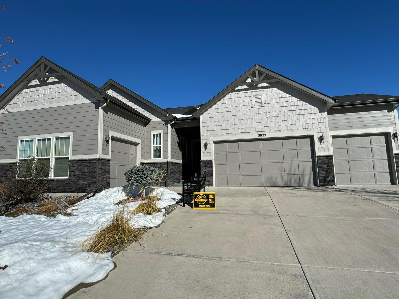 A one-story suburban home with a gray and stone facade, a multi-car garage, and a driveway with patches of snow.