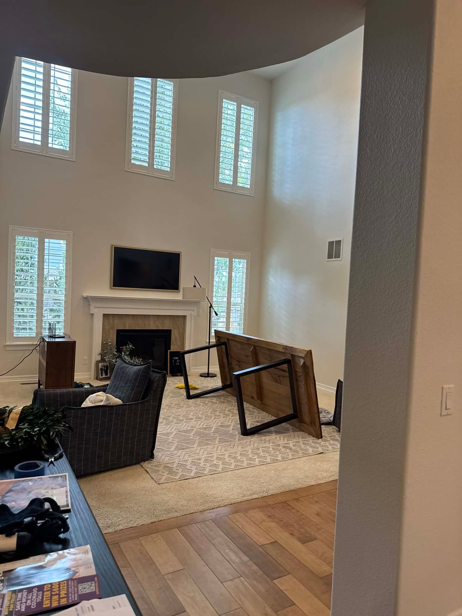 A high-ceiling living room with a fireplace, a TV, a gray armchair, and a wooden table overturned on a patterned rug.