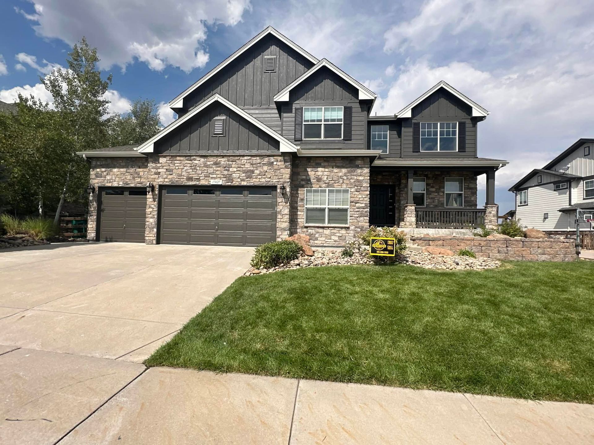 A two-story house with stone masonry, dark gray siding, a three-car garage, and a manicured lawn under a cloudy blue sky.
