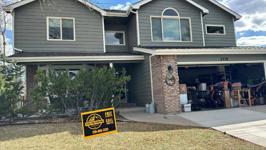 A two-story grey house with a brick pillar, open garage filled with storage, and a yellow real estate sign on the lawn.