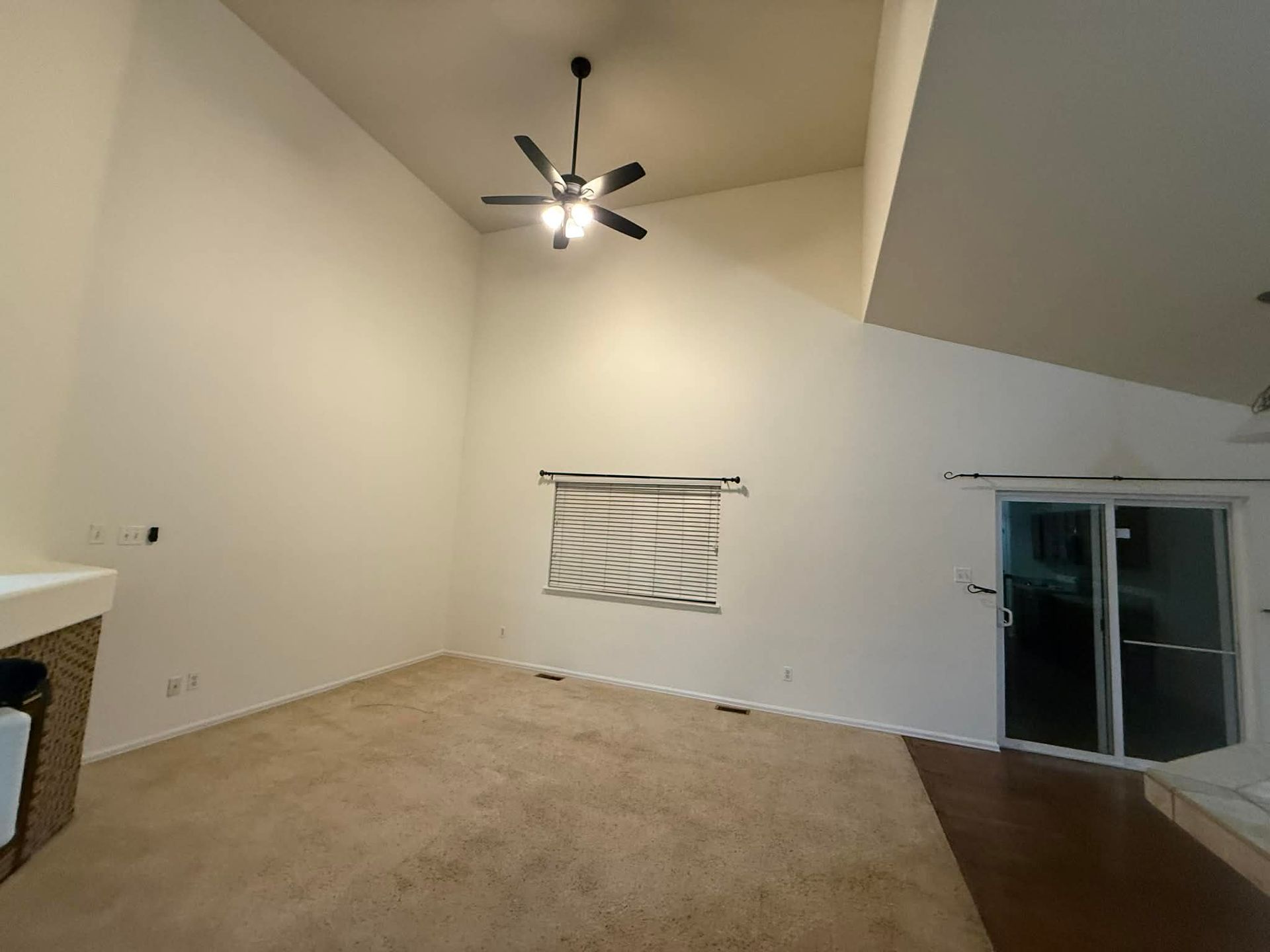 A spacious, empty room with high, angled ceilings, beige carpet, white walls, a ceiling fan, and sliding glass doors.