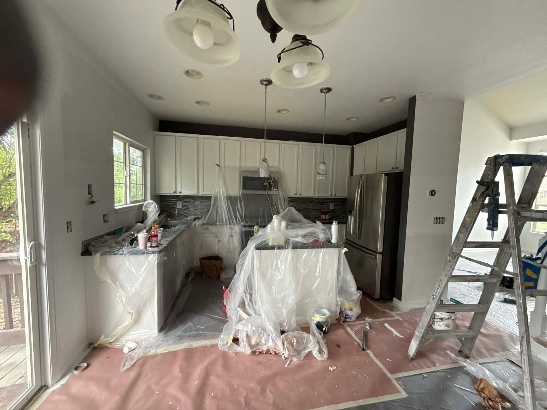 A kitchen undergoing renovation with white cabinets, stainless steel appliances, plastic sheeting, and a ladder.