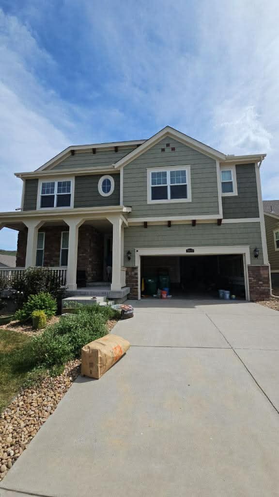 A two-story green suburban house with a two-car garage, front porch, stone accents, and a concrete driveway.