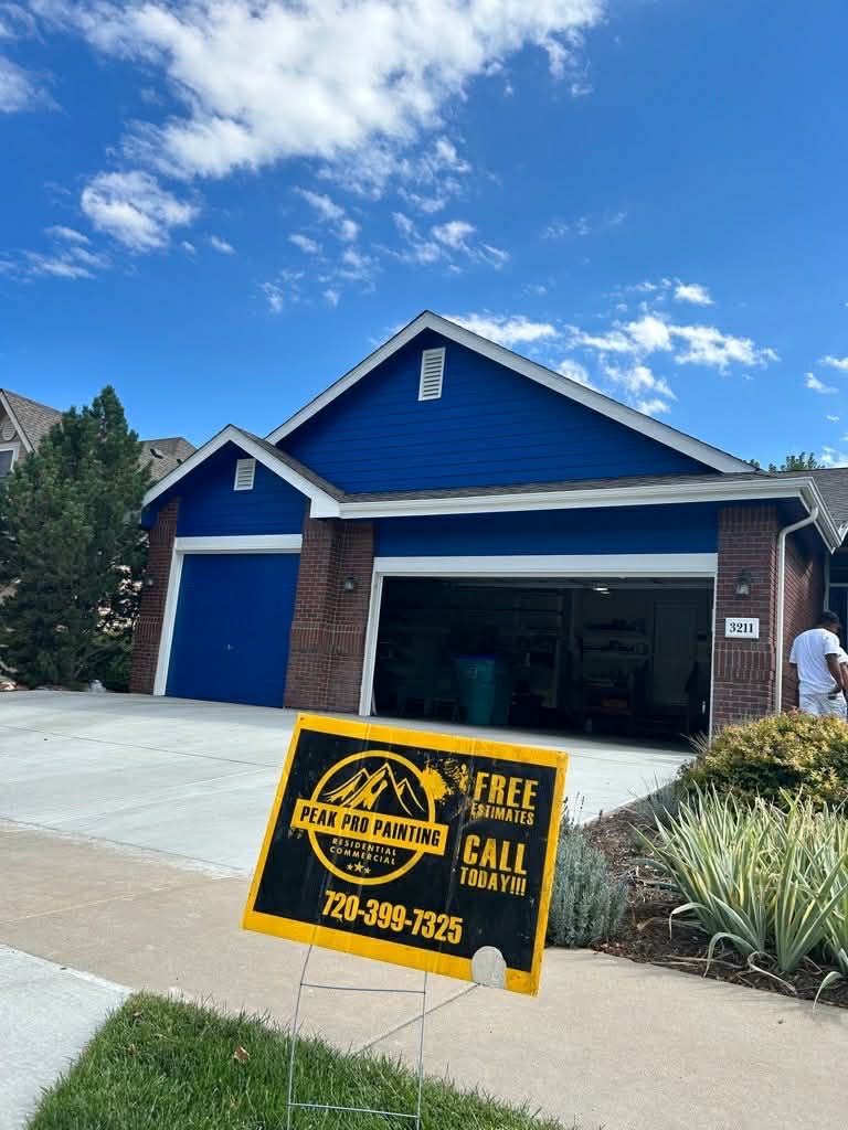 A blue-sided garage with a partially open door, with a yellow and black promotional sign in the foreground on a sidewalk.