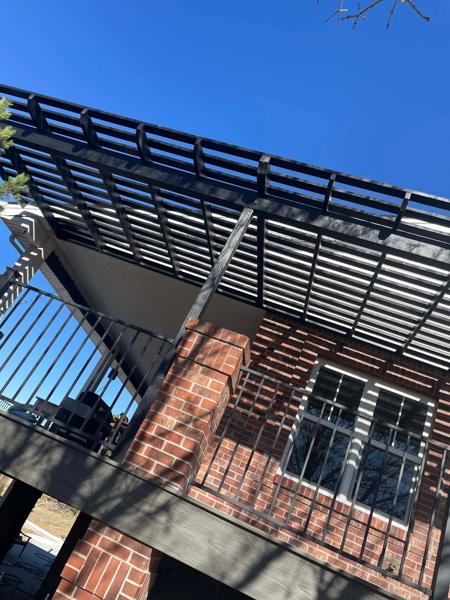 A low-angle view of a brick house with a black metal balcony railing and a slatted pergola roof against a clear blue sky.