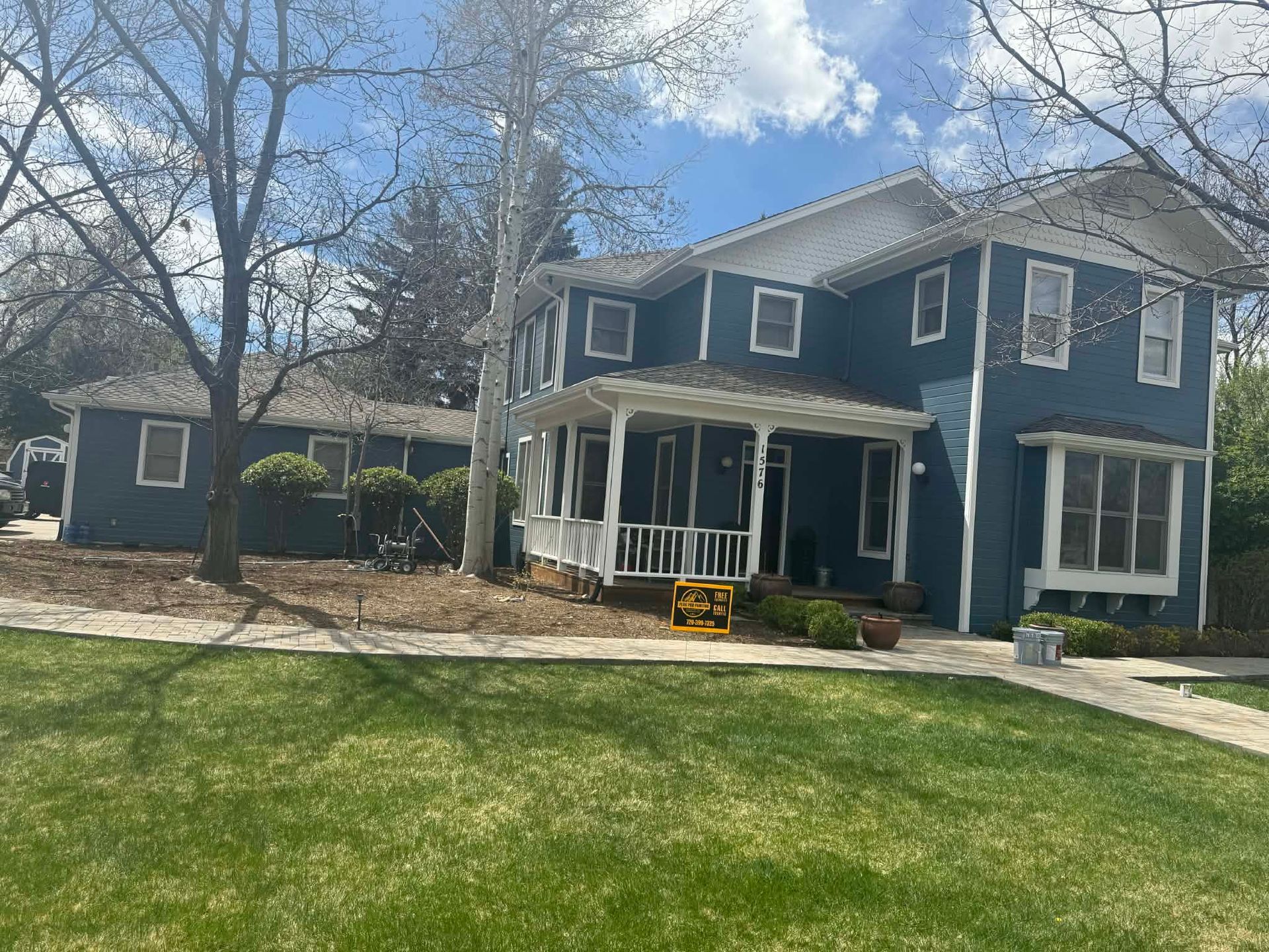 A two-story blue house with a white front porch and an attached garage, set on a green lawn under a sunny sky.