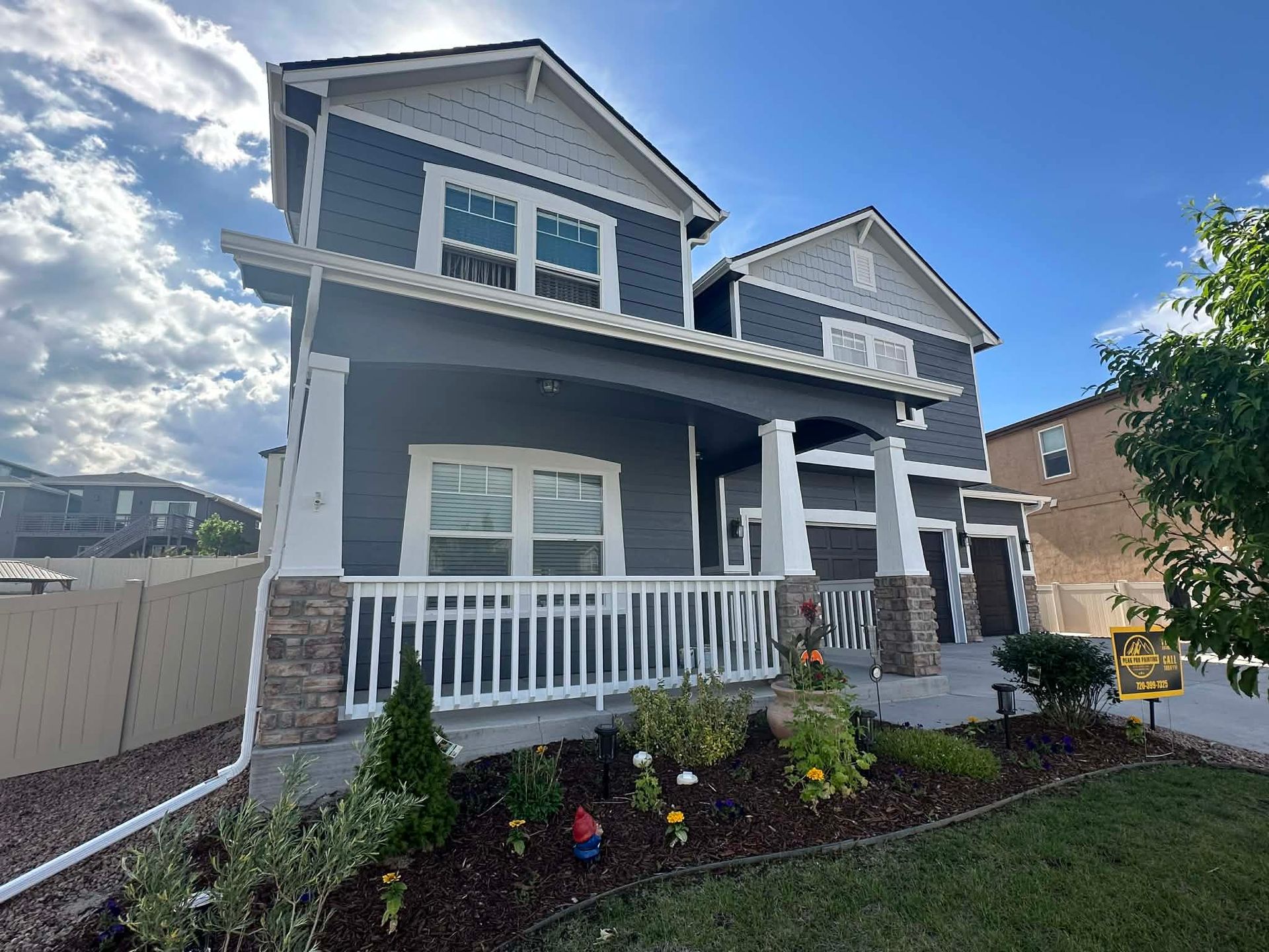 A two-story house with dark grey siding, white trim, a front porch, and stone accents, set against a sunny blue sky.