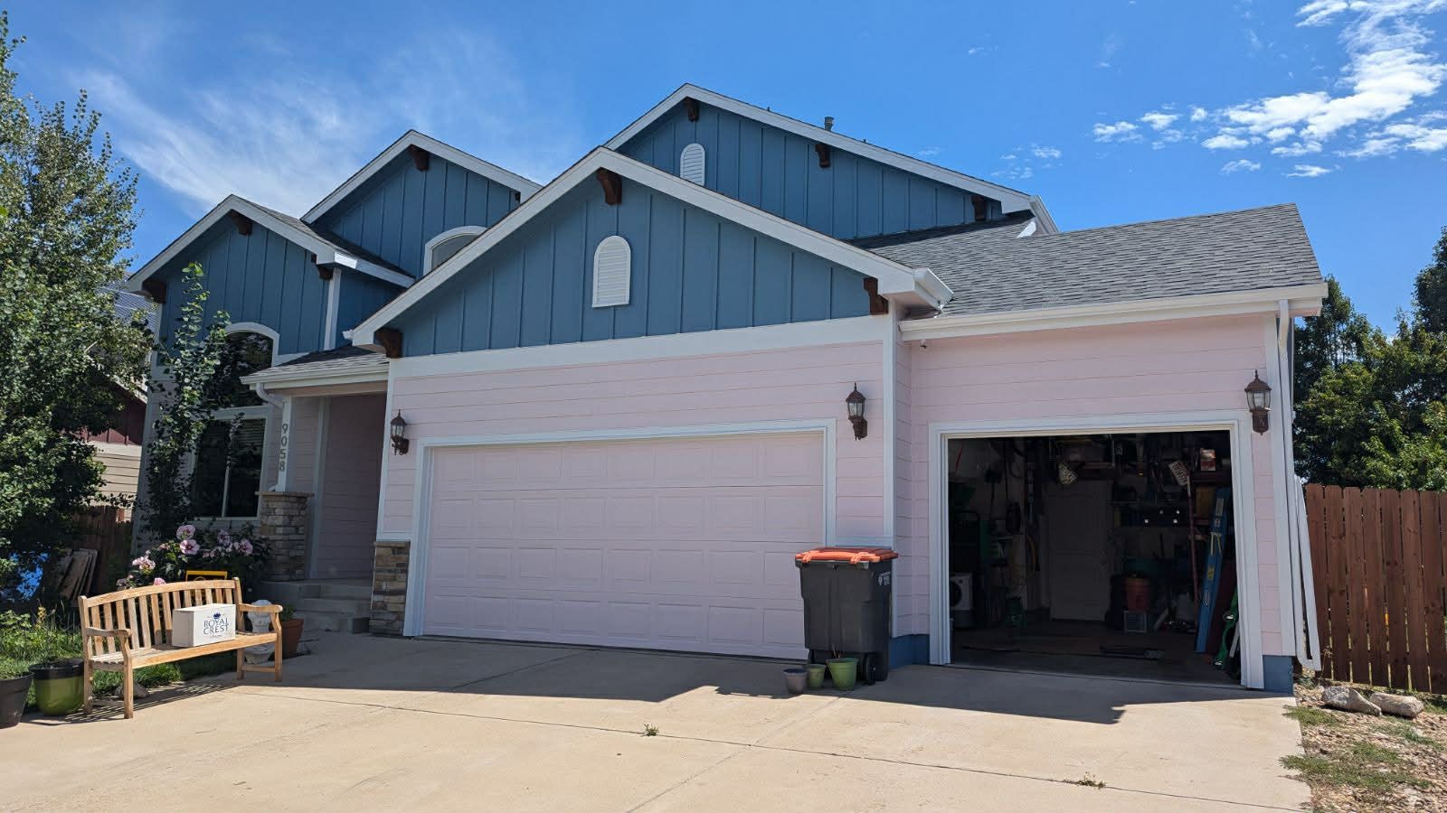 A suburban house with pink stucco walls, blue vertical siding on the gables, a two-car garage, and a wooden bench.