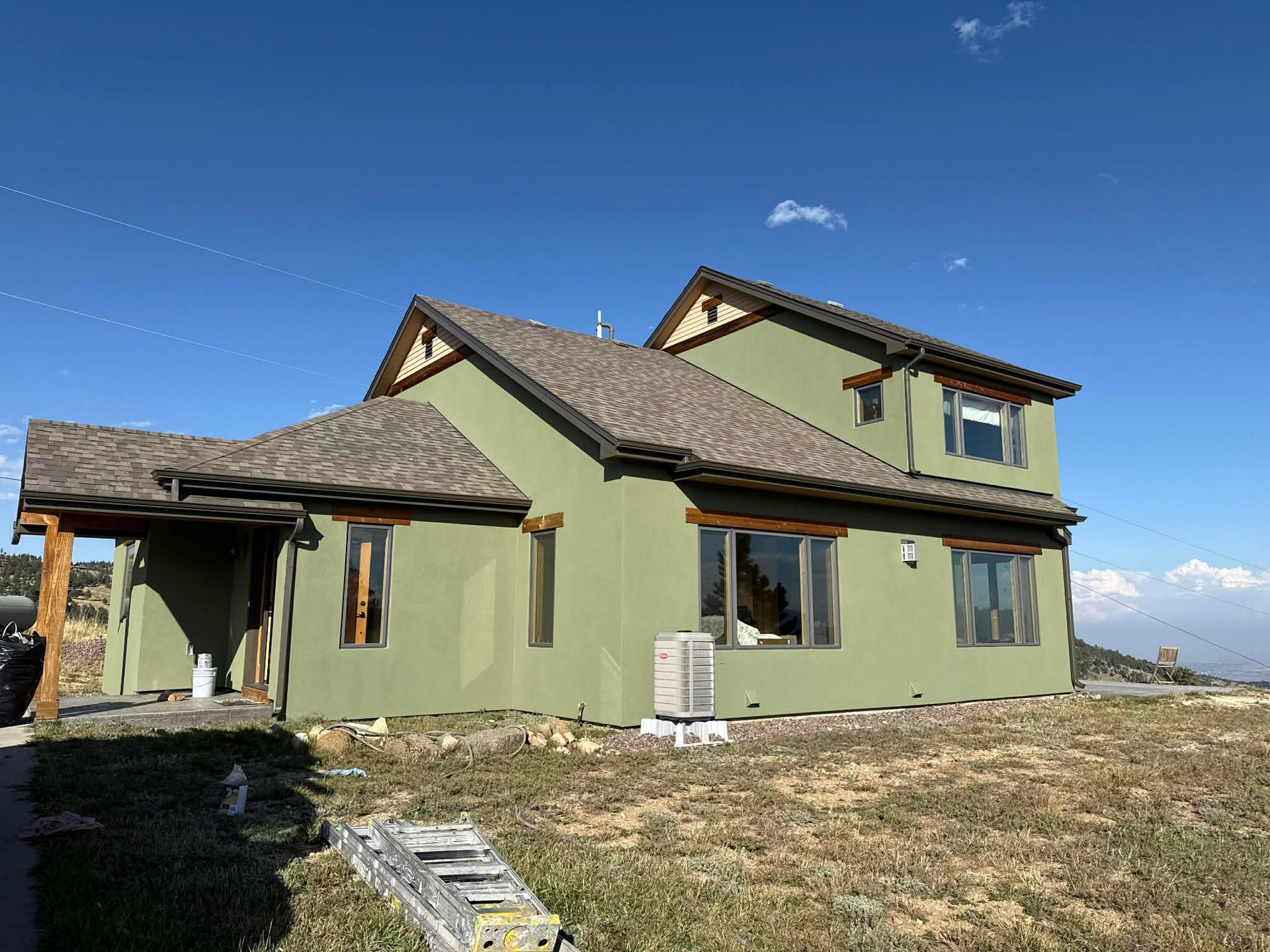 A two-story, olive-green house with a brown roof sits on a dry, grassy landscape under a clear blue sky.
