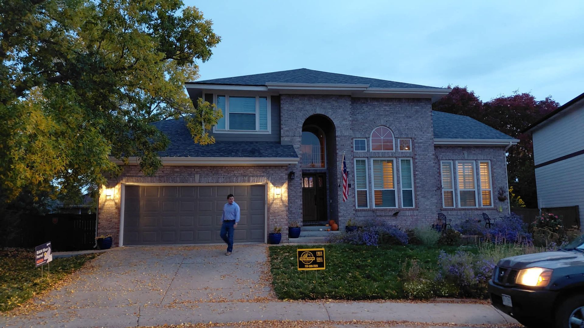 A person stands on the driveway of a two-story brick home at dusk, with a dark car parked on the right.