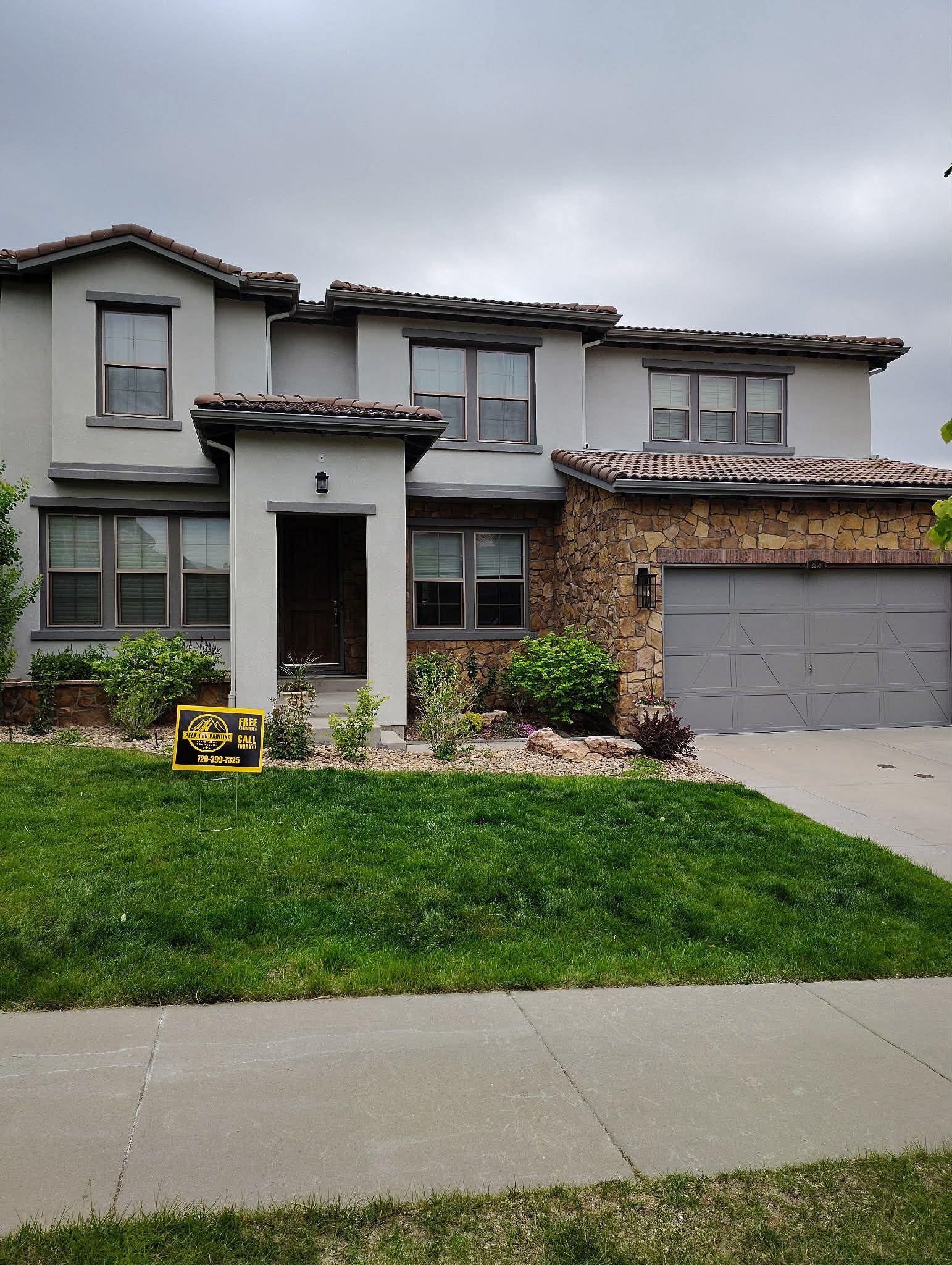 A two-story suburban house with a gray exterior, stone accents on the garage side, and a tile roof under a cloudy sky.