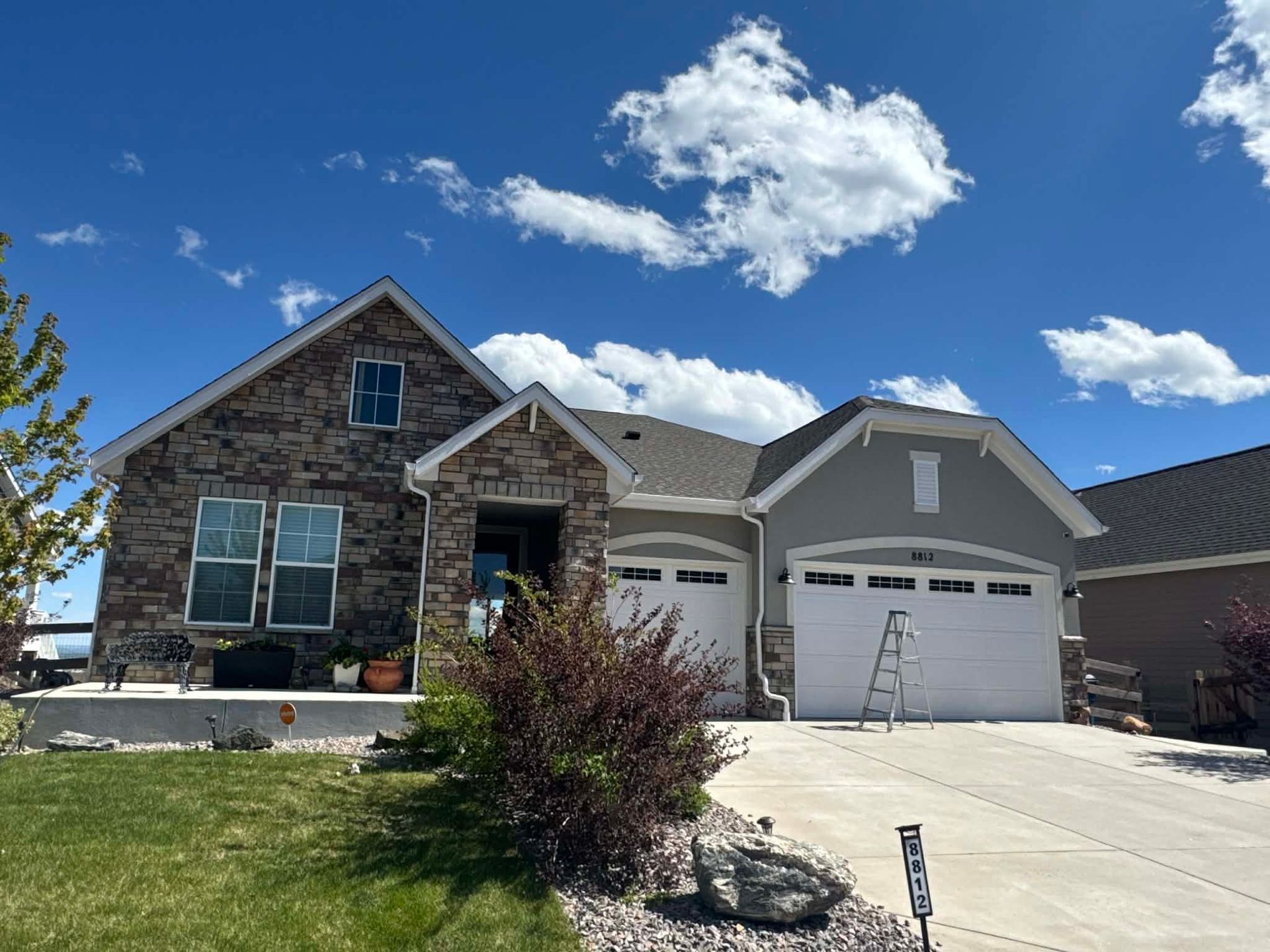 A single-story suburban house with stone and gray siding, a two-car garage, a concrete driveway, and a blue, cloudy sky.