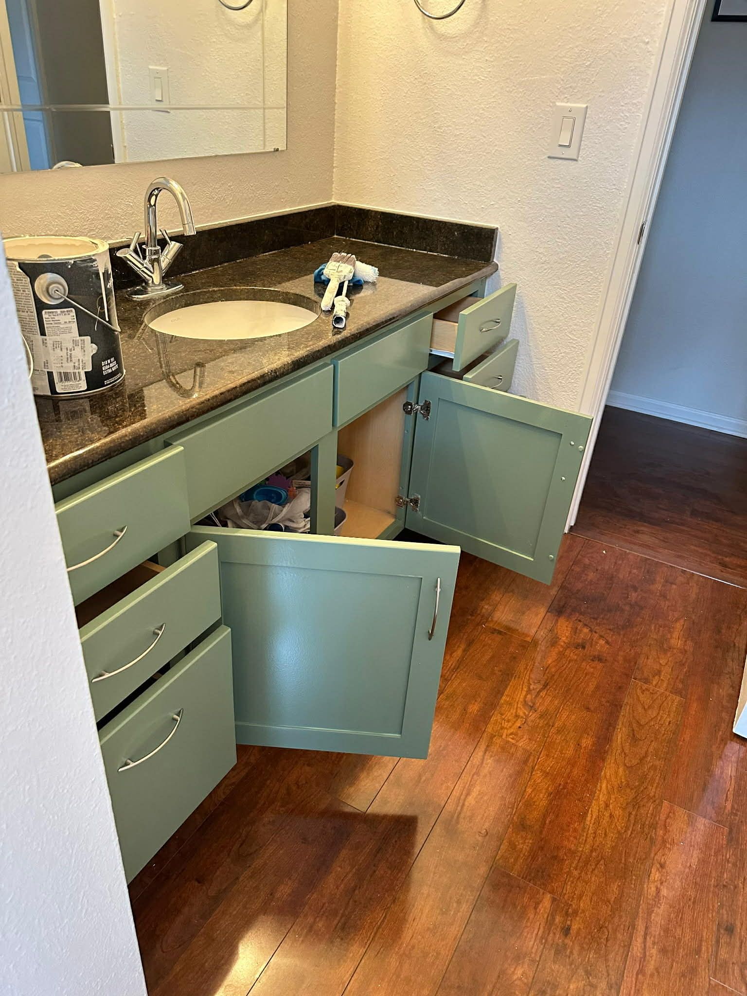 A sage green bathroom vanity with open doors and drawers, situated on a dark wood floor in a bathroom with textured walls.