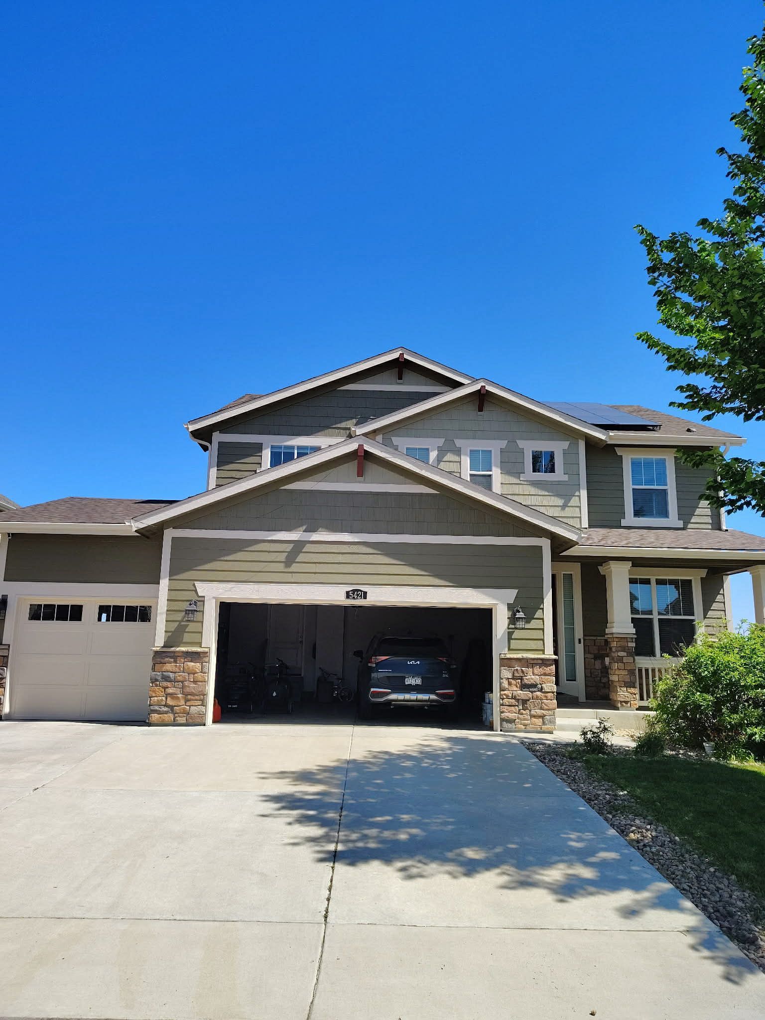 A two-story suburban house with olive green siding, stone accents, a three-car garage, and a concrete driveway.