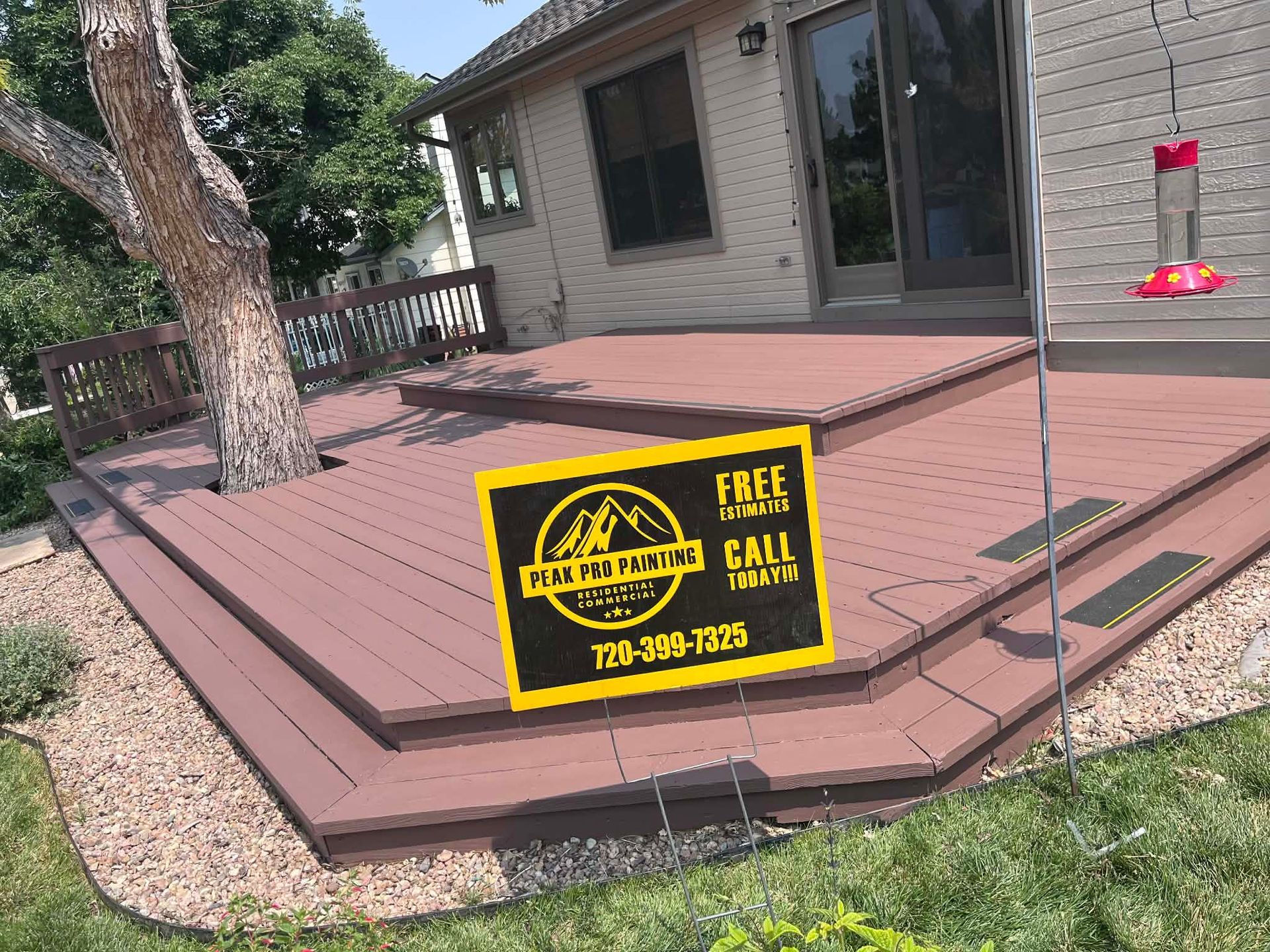 A brown composite deck with steps wraps around a tree, featuring a yellow advertisement sign in the foreground.