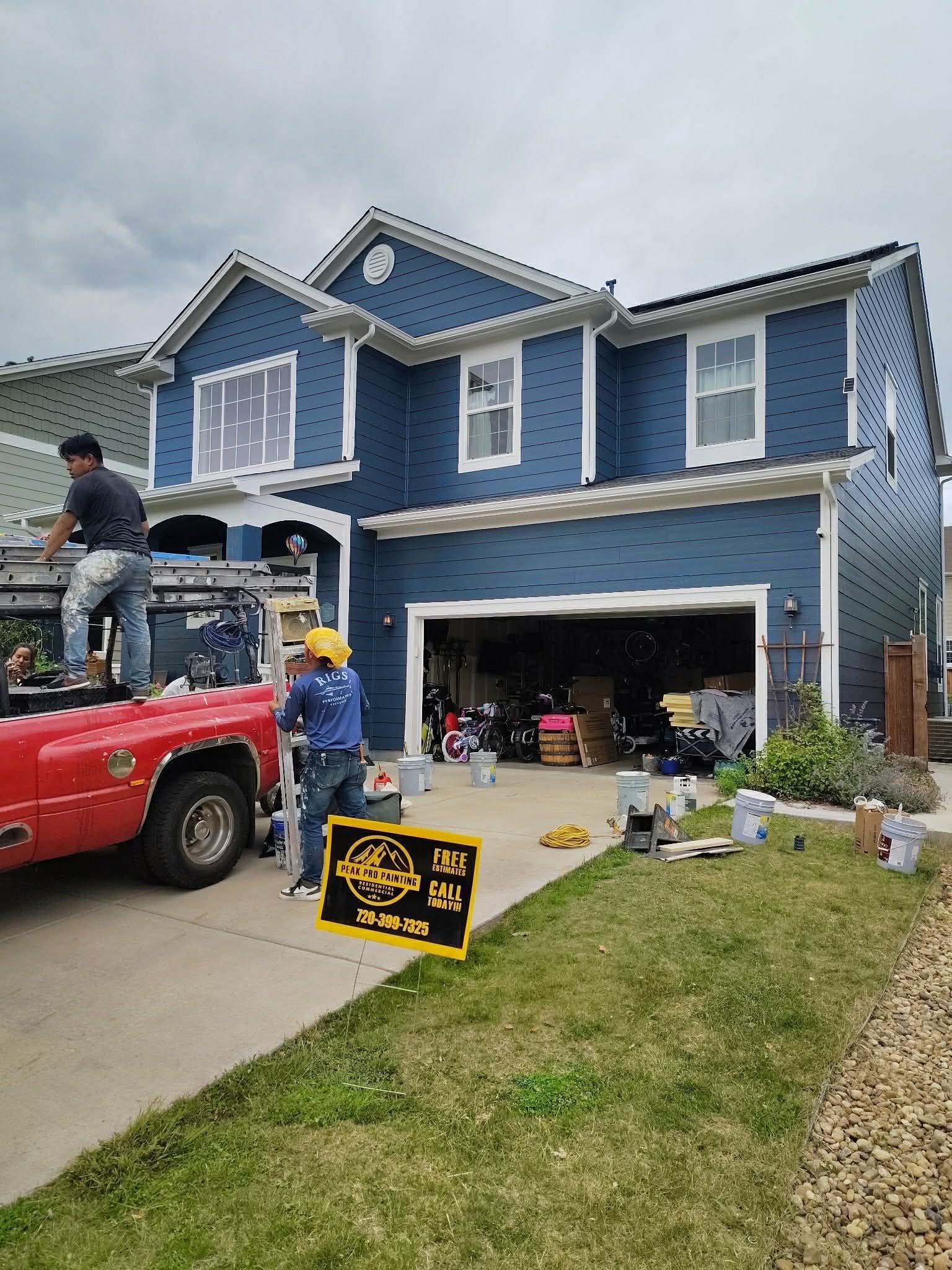 Workers renovate the exterior of a dark blue two-story house with white trim, parked in front of a red truck.