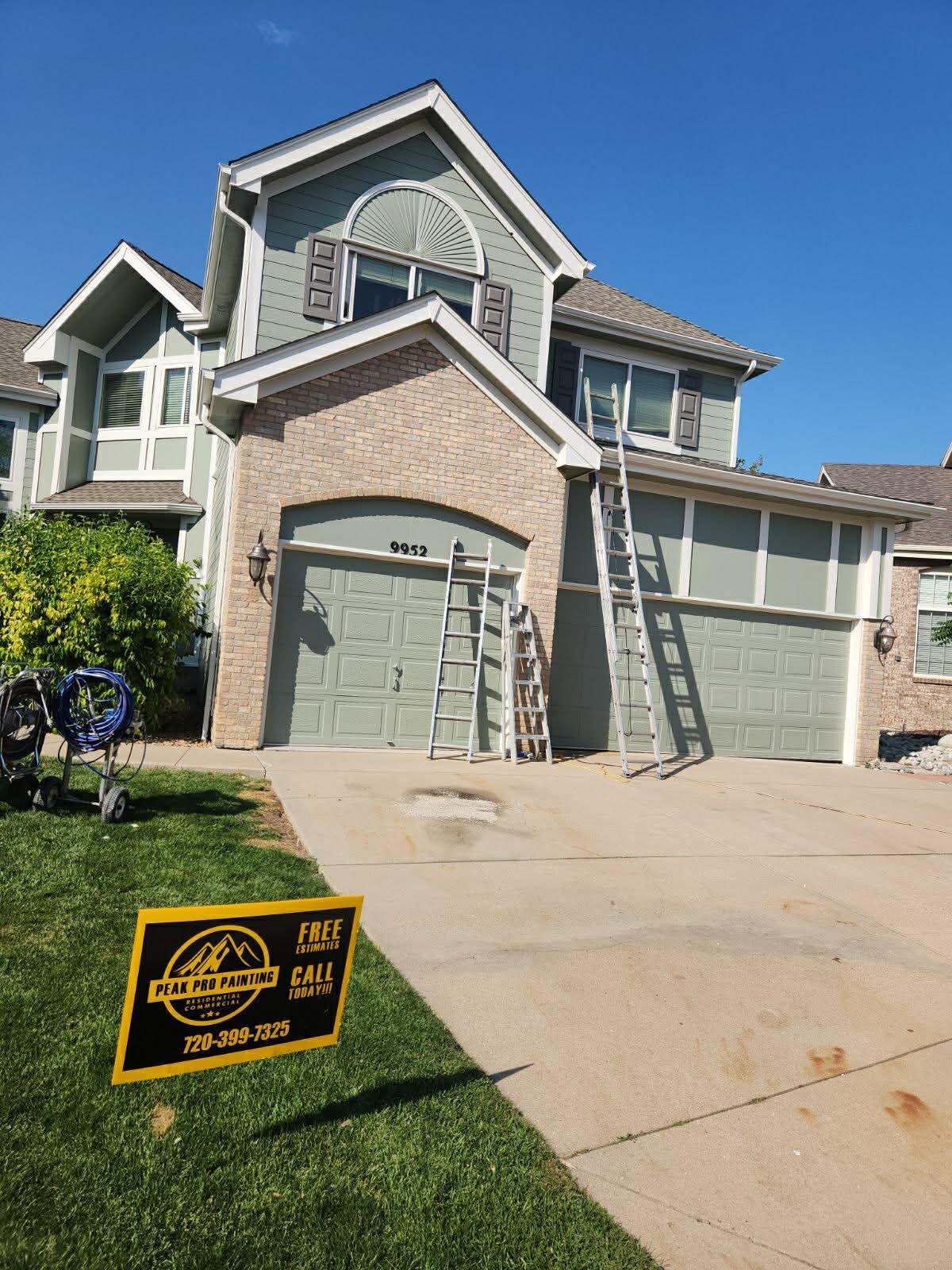 A two-story tan and sage green suburban home with two ladders leaning against the exterior and a yard sign in the grass.