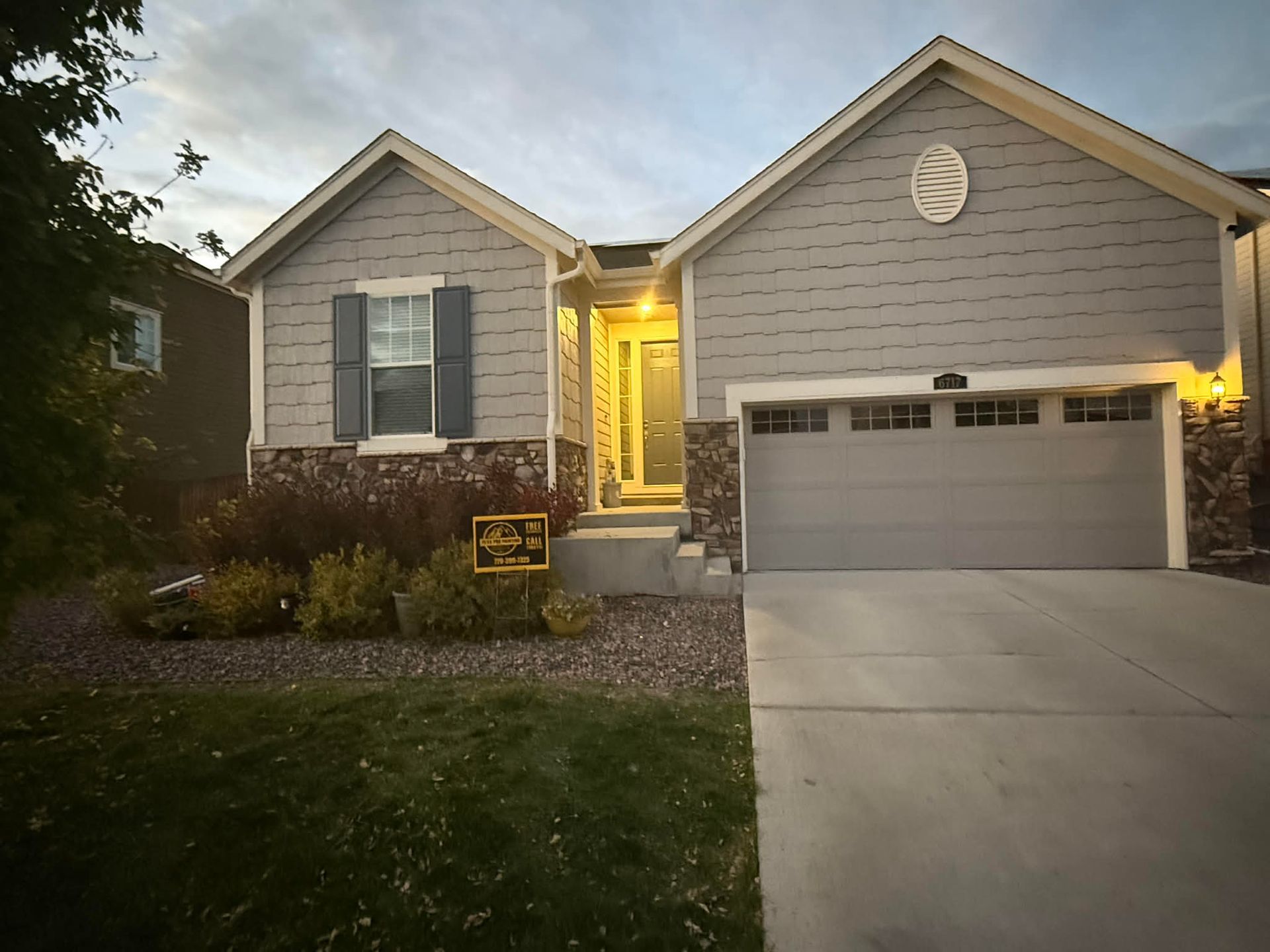 A single-story suburban house with grey siding, stone accents, a two-car garage, and a light-filled entryway at dusk.