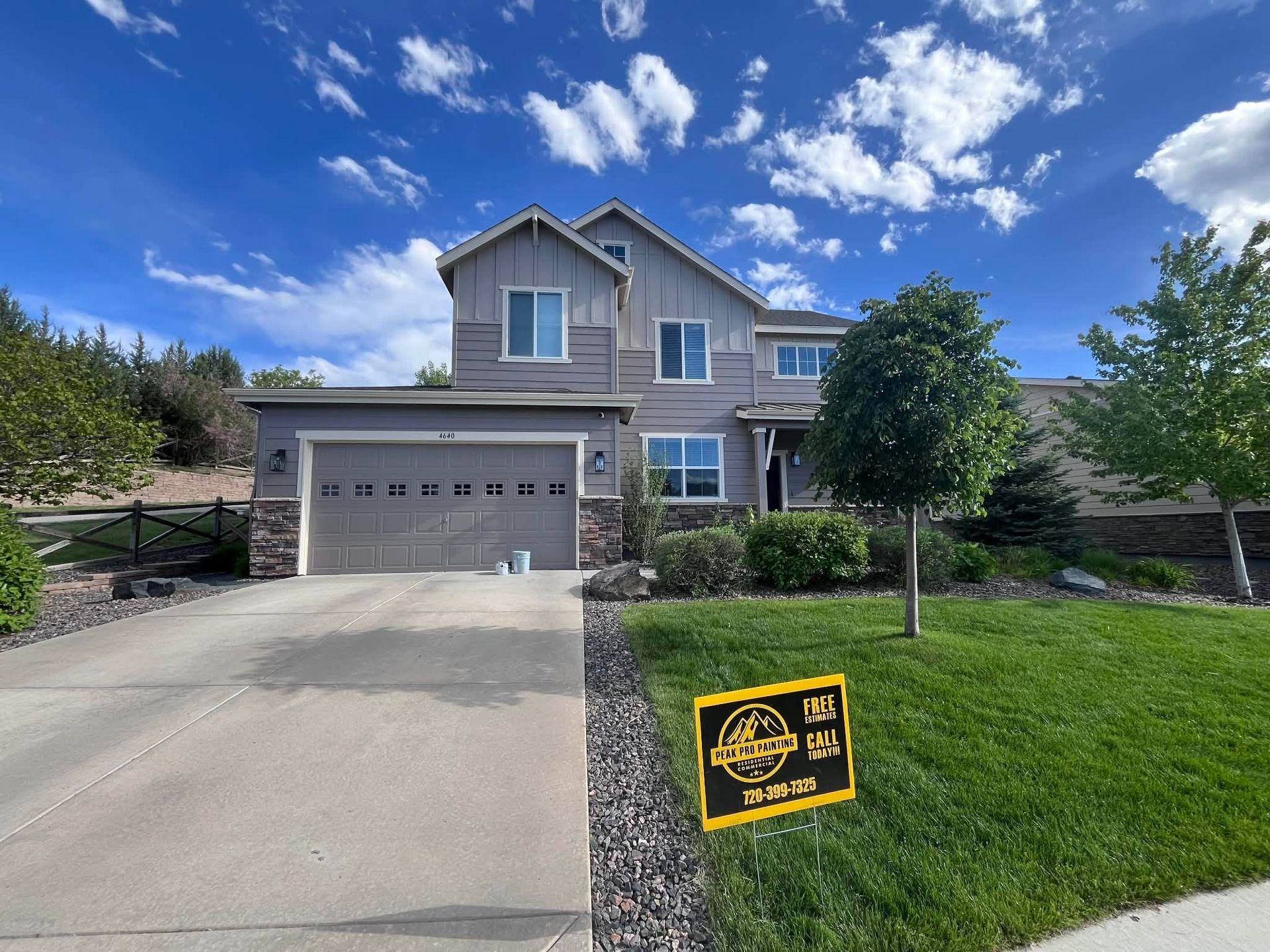 A two-story grey suburban house with a garage, driveway, and front lawn under a blue sky with scattered clouds.