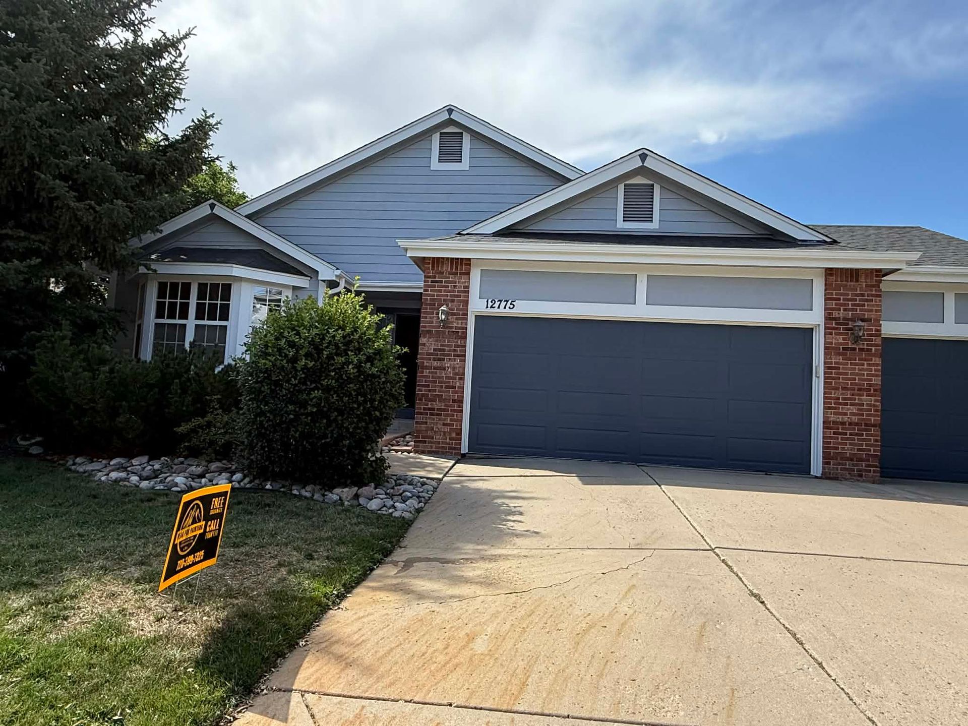 A one-story suburban home with blue siding, brick accents, a gray garage door, and a yellow sign on the lawn.