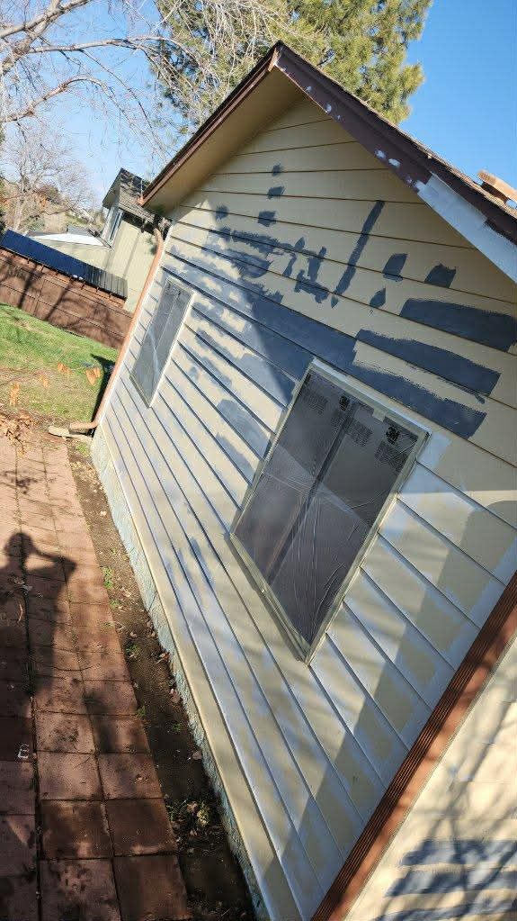 A side view of a house exterior with tan horizontal siding featuring dark gray paint stripes being applied.
