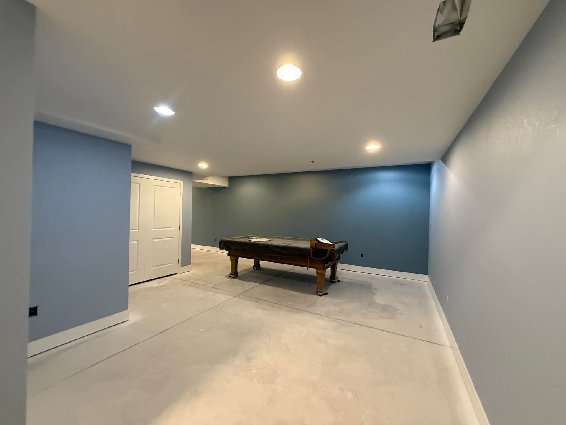 A pool table sits in the center of an empty basement room with light blue walls and recessed lighting.
