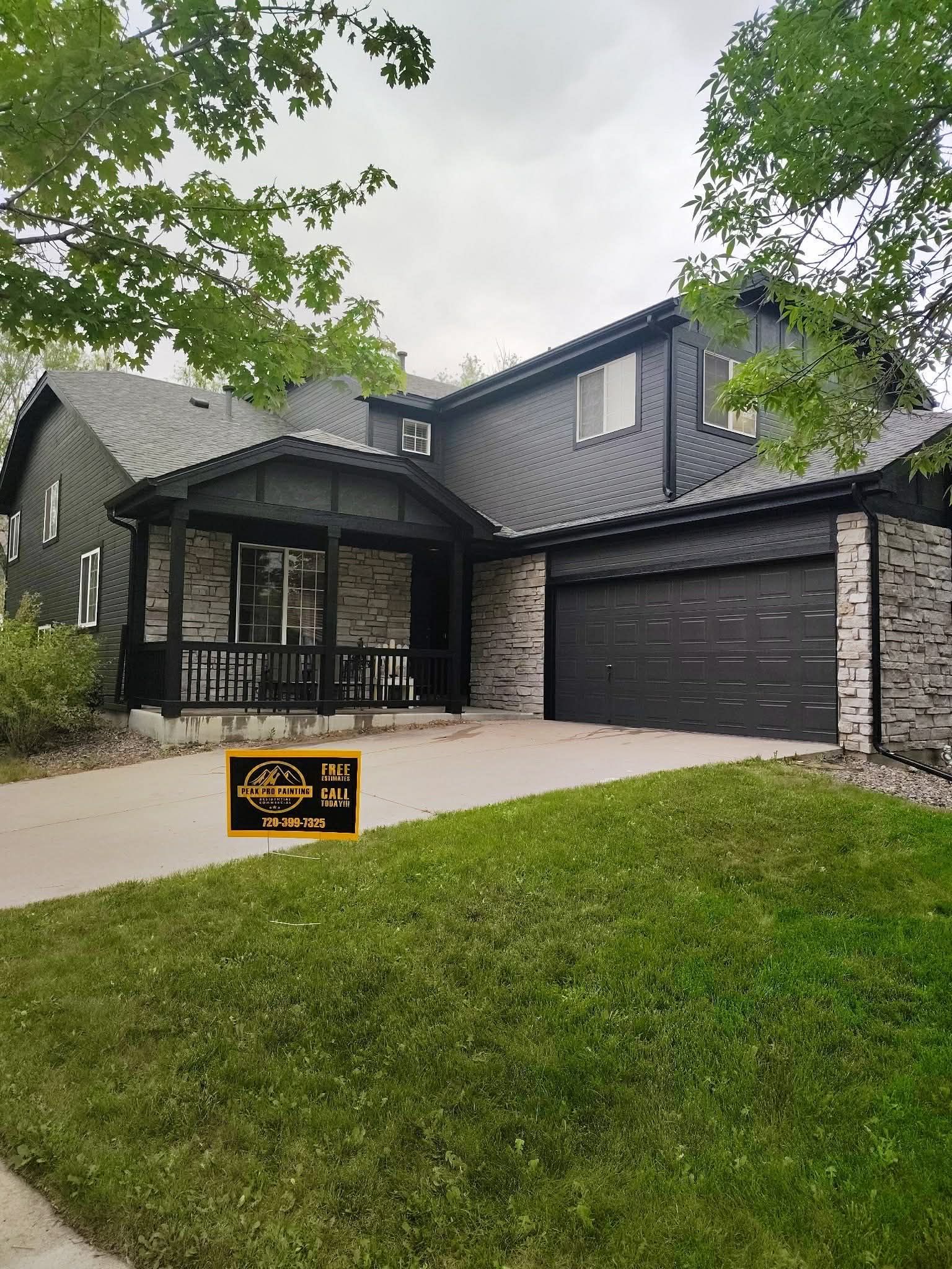 A two-story house with dark siding and stone veneer, featuring a front porch and an attached garage under a cloudy sky.