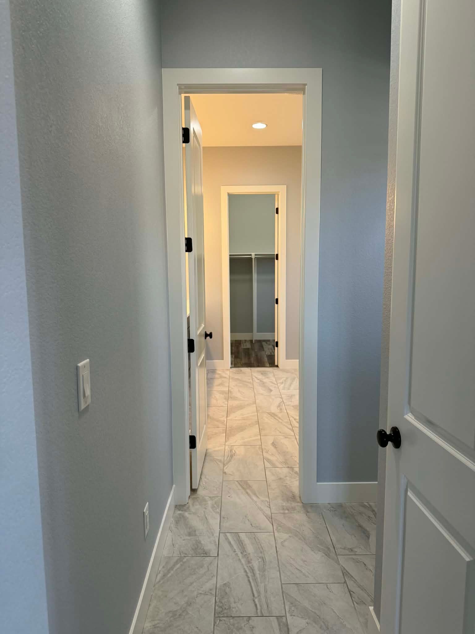 A hallway with gray walls and light wood-look tile flooring leads through an open door into a room with a closet.