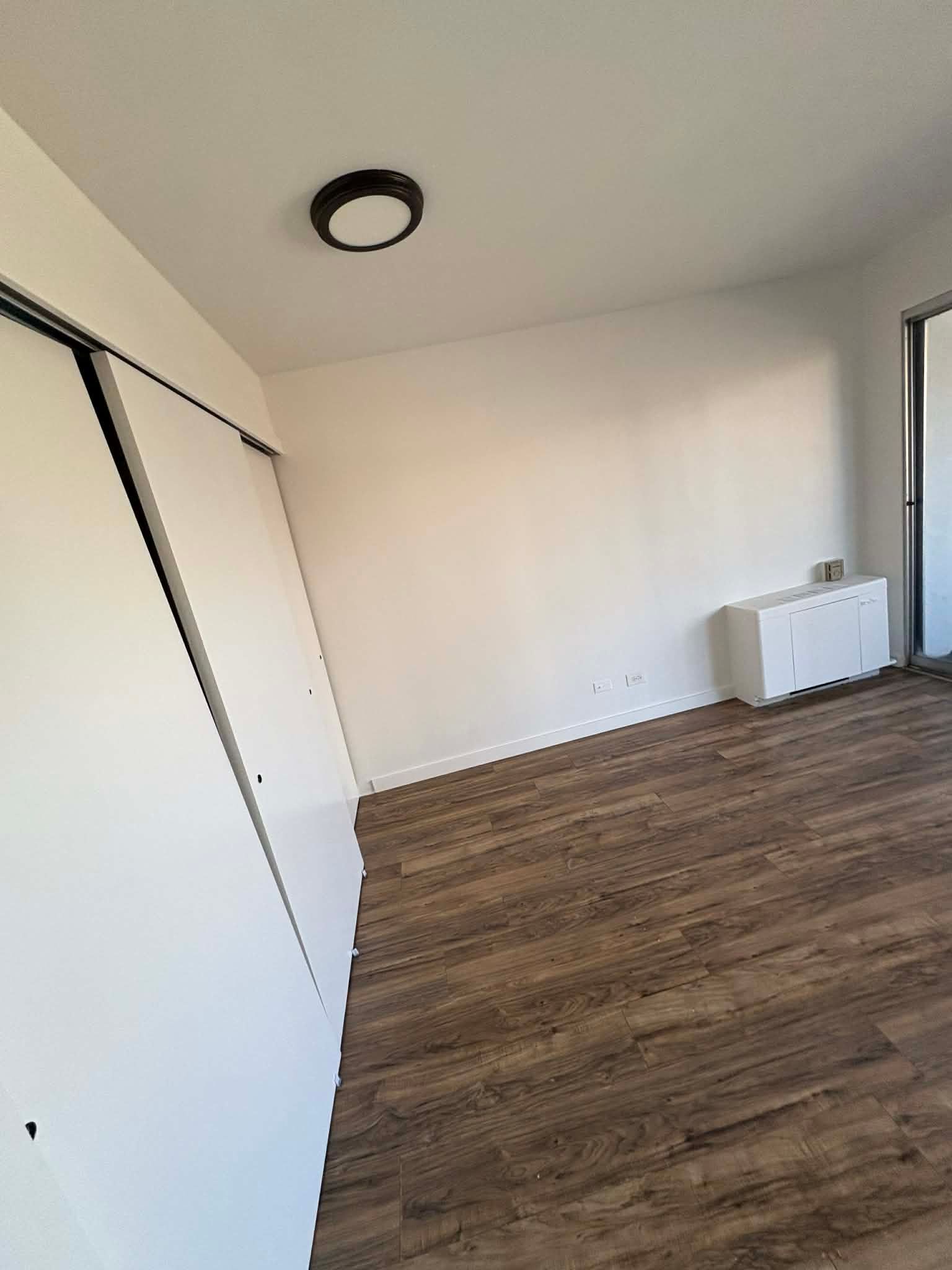 An empty room featuring light-colored walls, wood-grain flooring, white sliding closet doors, and a ceiling light.