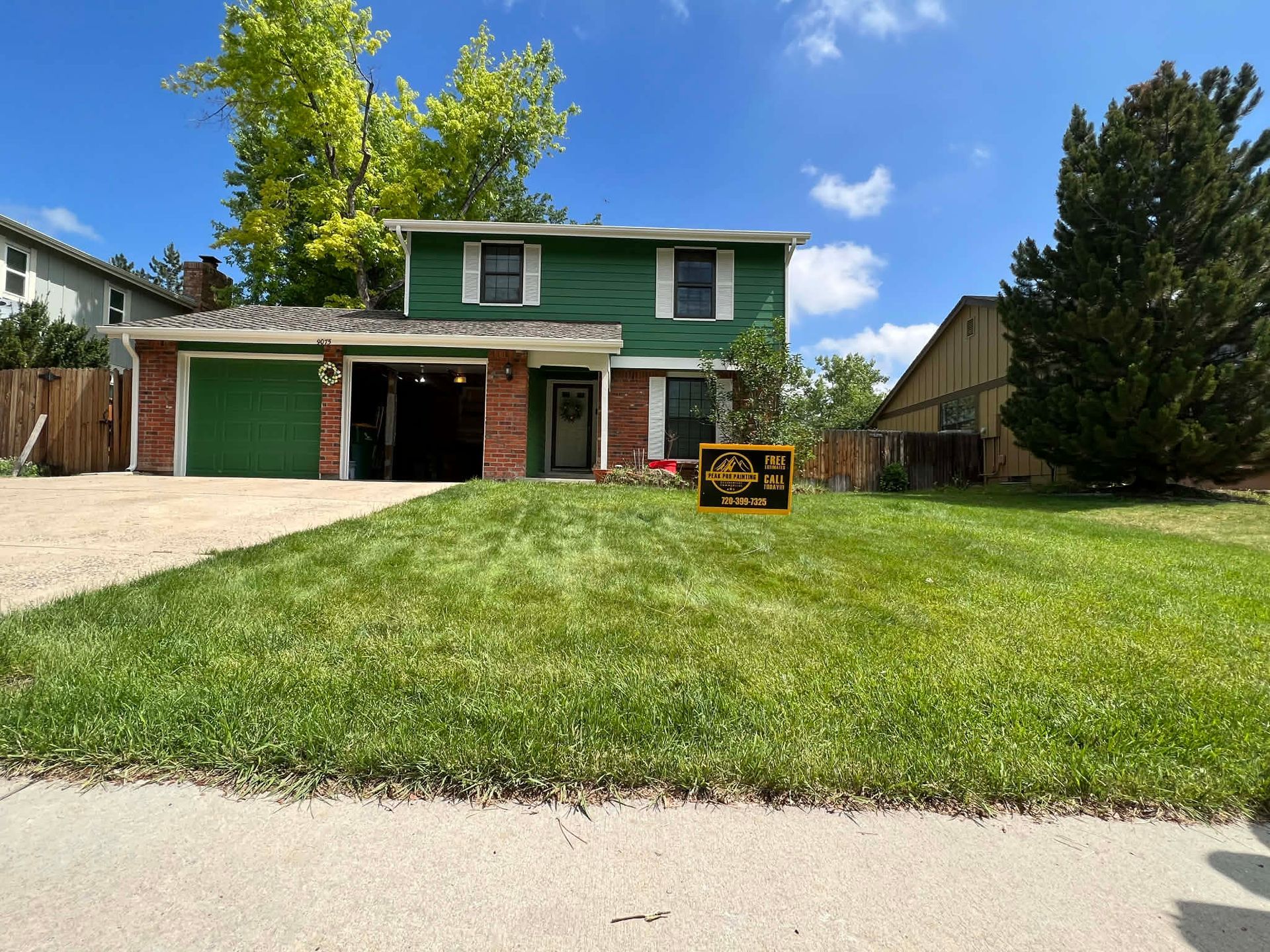 A two-story green house with a brick entryway and a two-car garage sits behind a large, well-manicured lawn on a sunny day.
