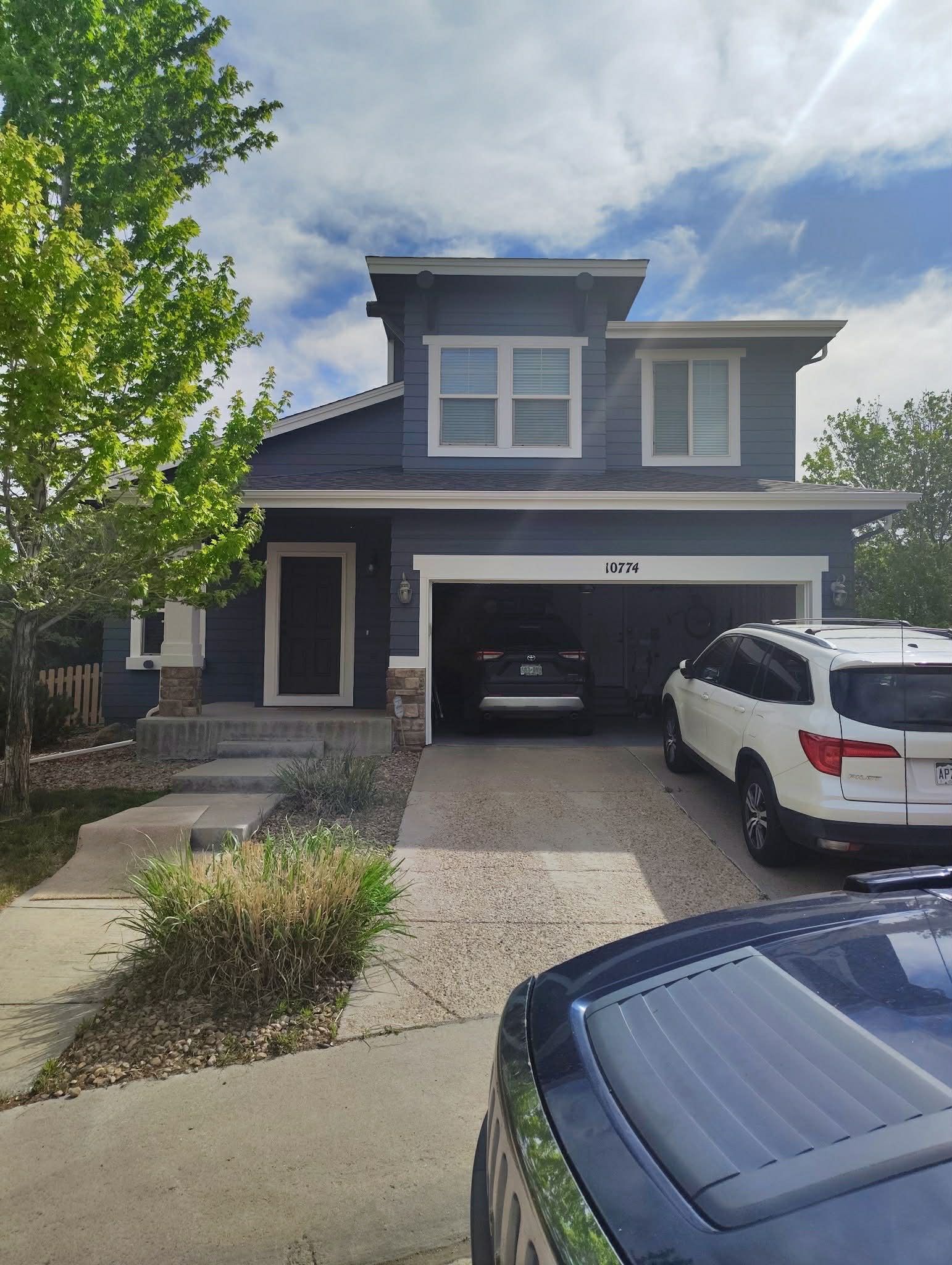 A two-story blue house with stone accents, a garage, a driveway with a white SUV, and a small front lawn under a cloudy sky.