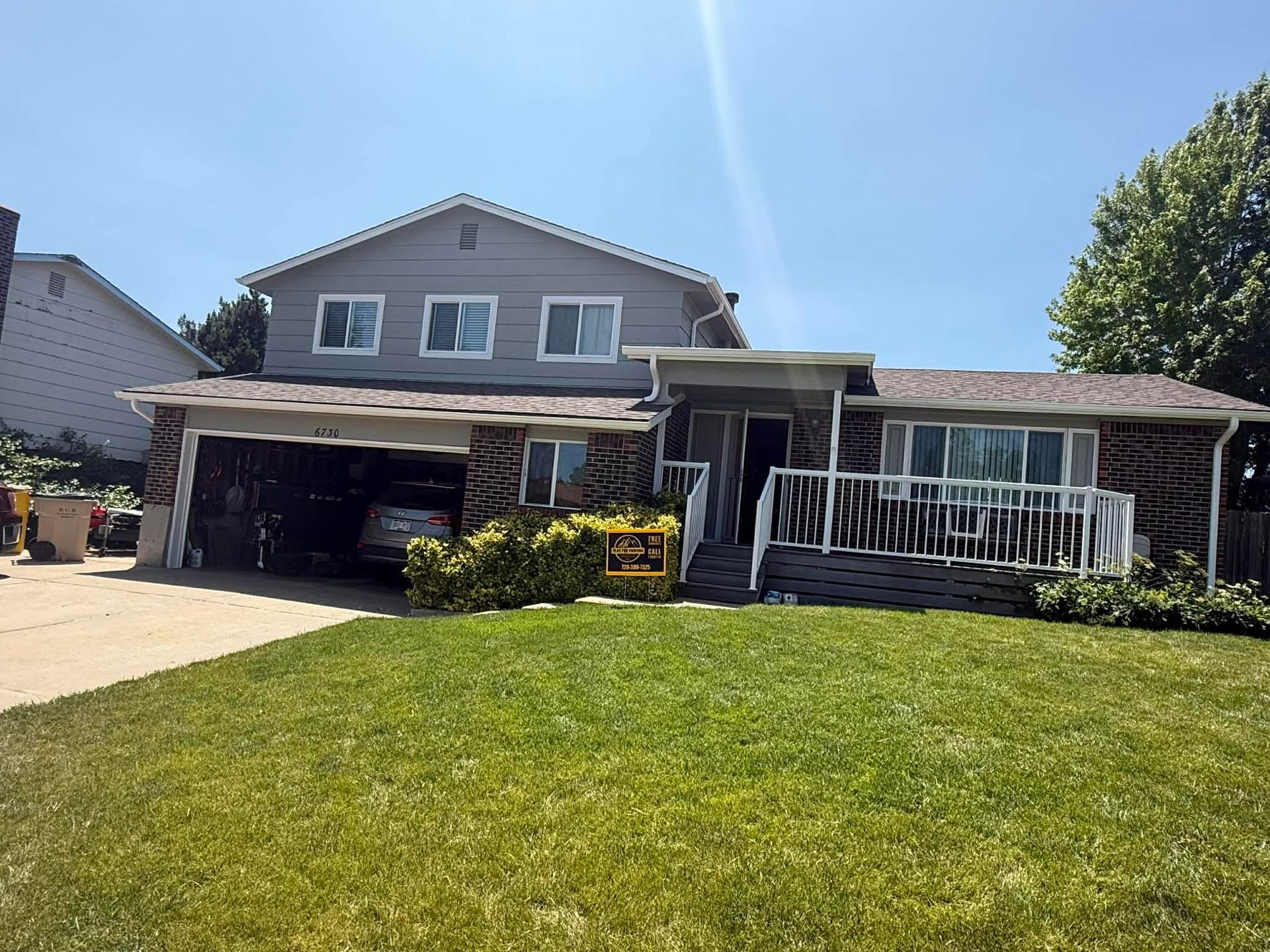 A grey two-story house with a brick lower level, an attached garage, and a front porch with a white railing.