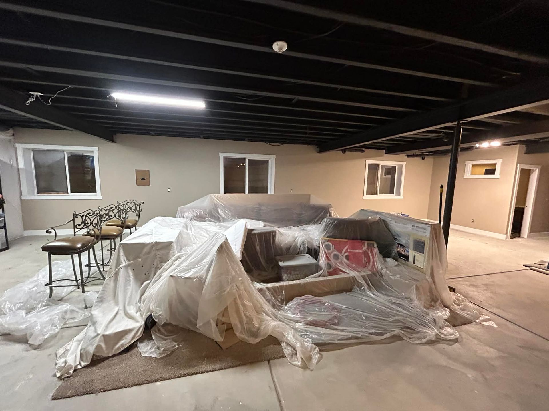Basement with a black ceiling, taupe walls, three windows, bar stools, and furniture covered in plastic sheeting.