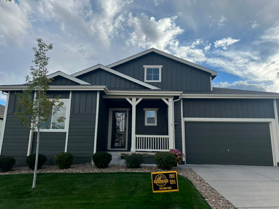 A modern dark gray house with a white front porch, a two-car garage, and a lawn with a yellow real estate sign.