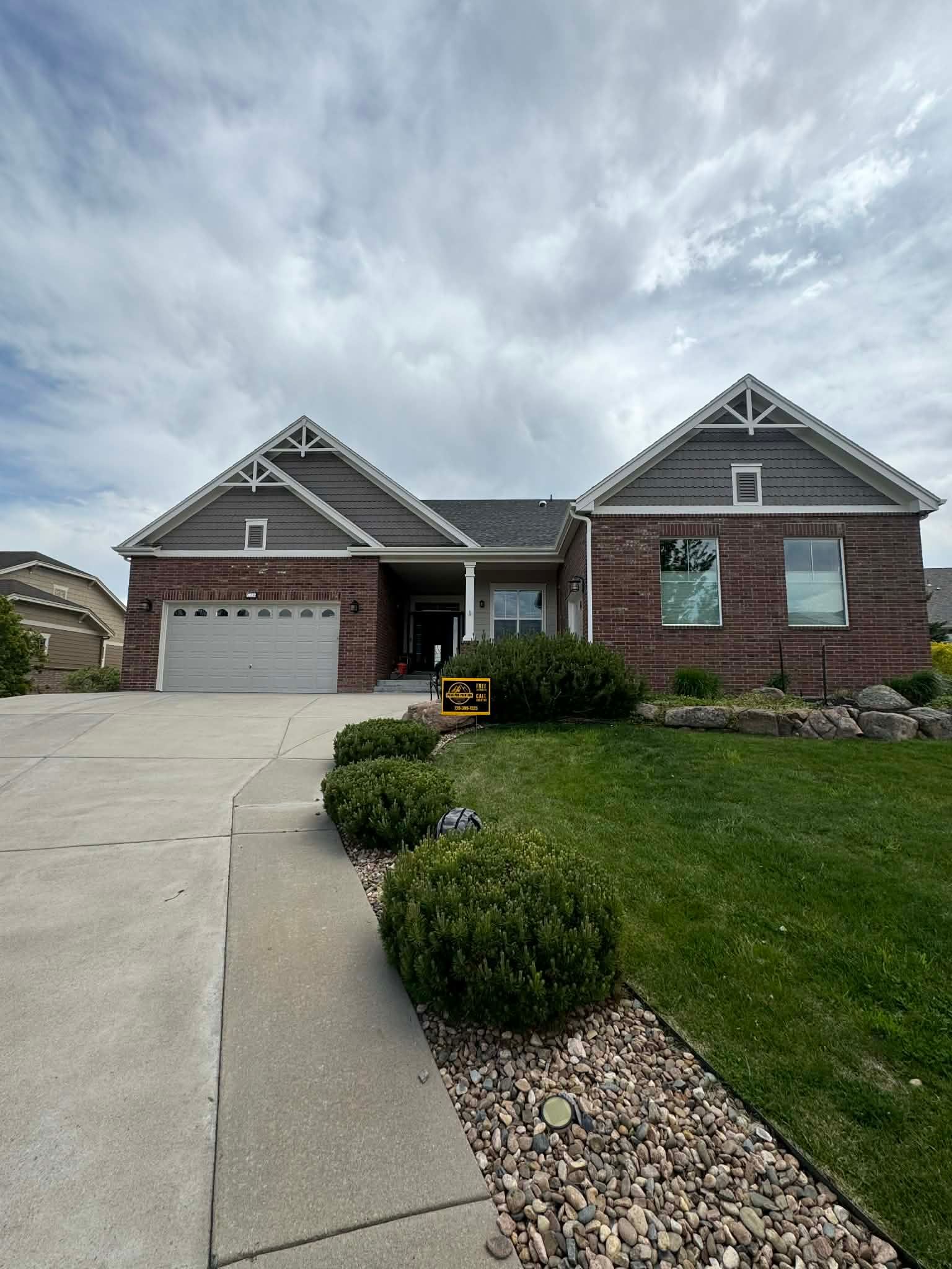 A single-story brick house with a two-car garage, grey gabled roofs, and a landscaped front lawn under a cloudy sky.